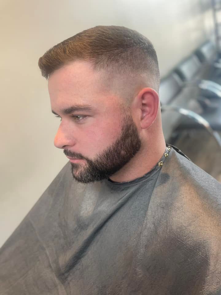 Man with a neatly faded haircut and full beard sitting in a barber chair, wearing a gray cape.