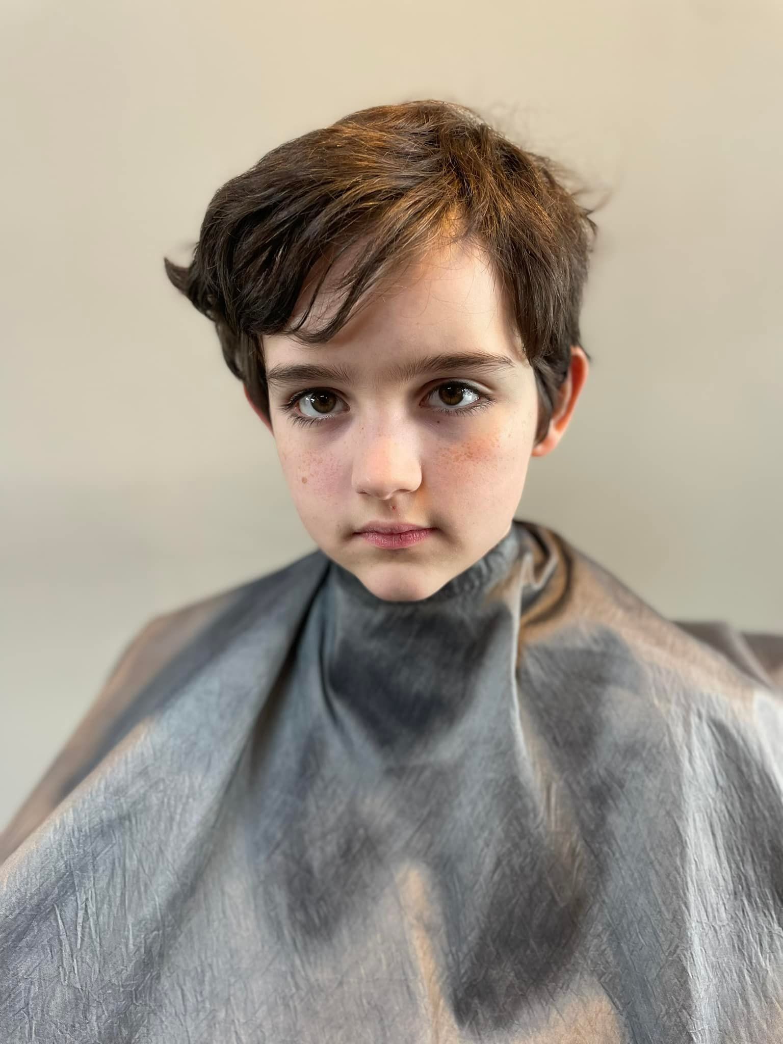 Child with short dark hair and a neutral expression, seated against a plain background wearing a gray salon cape.