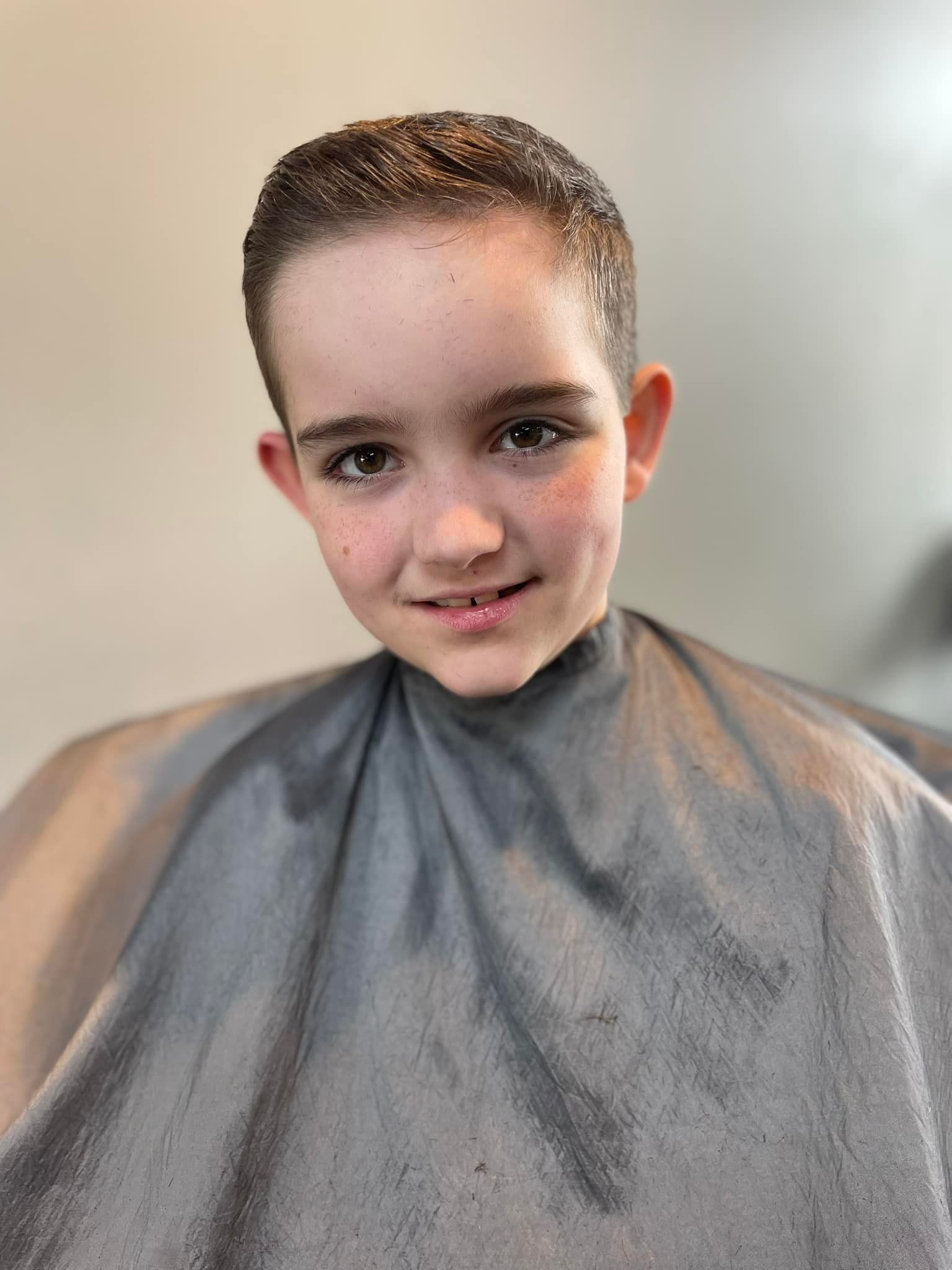 Smiling child in a gray barber cape, seated indoors with a fresh haircut