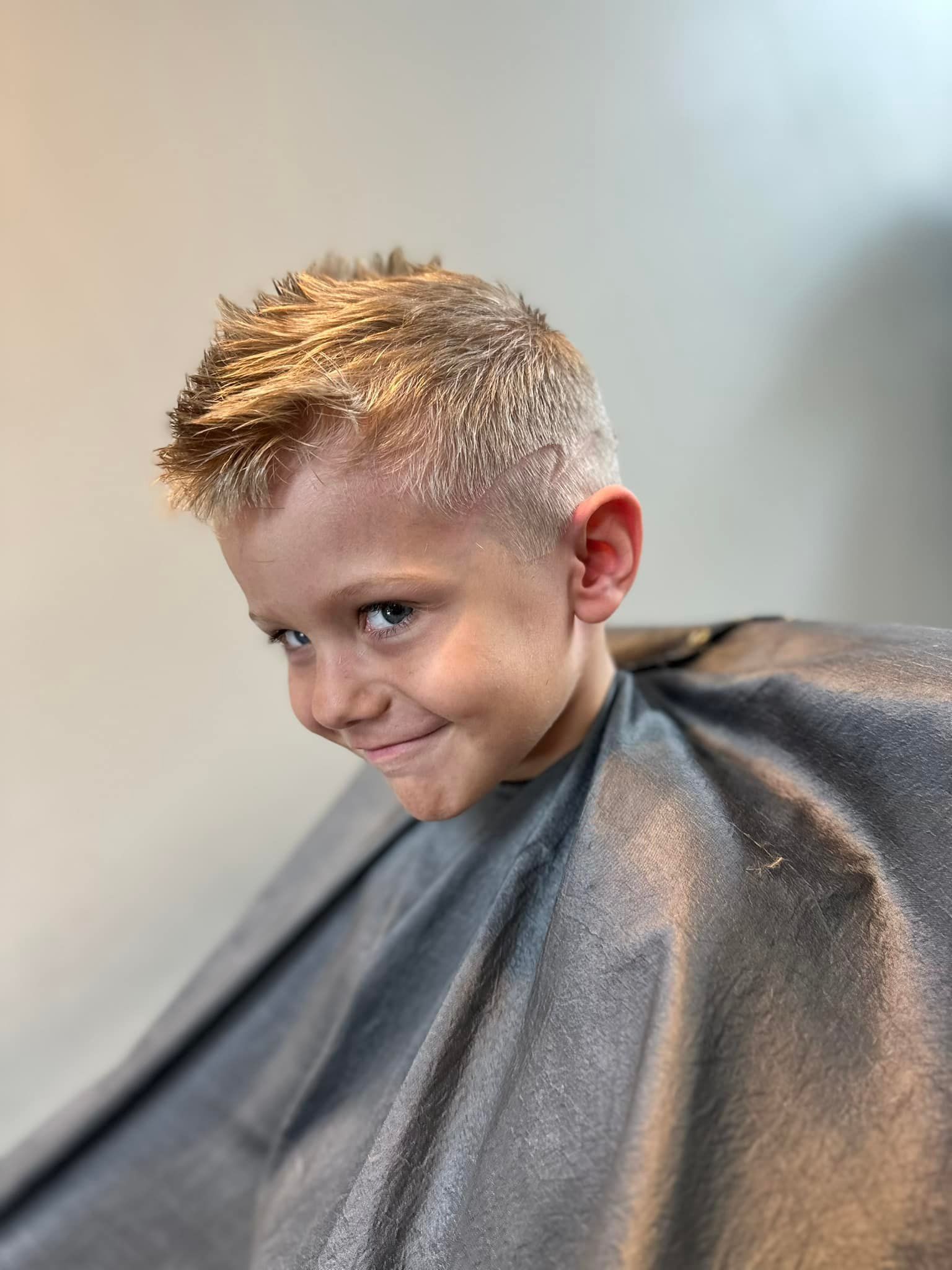 Child with short blond hair smiling under a gray haircut cape in a salon