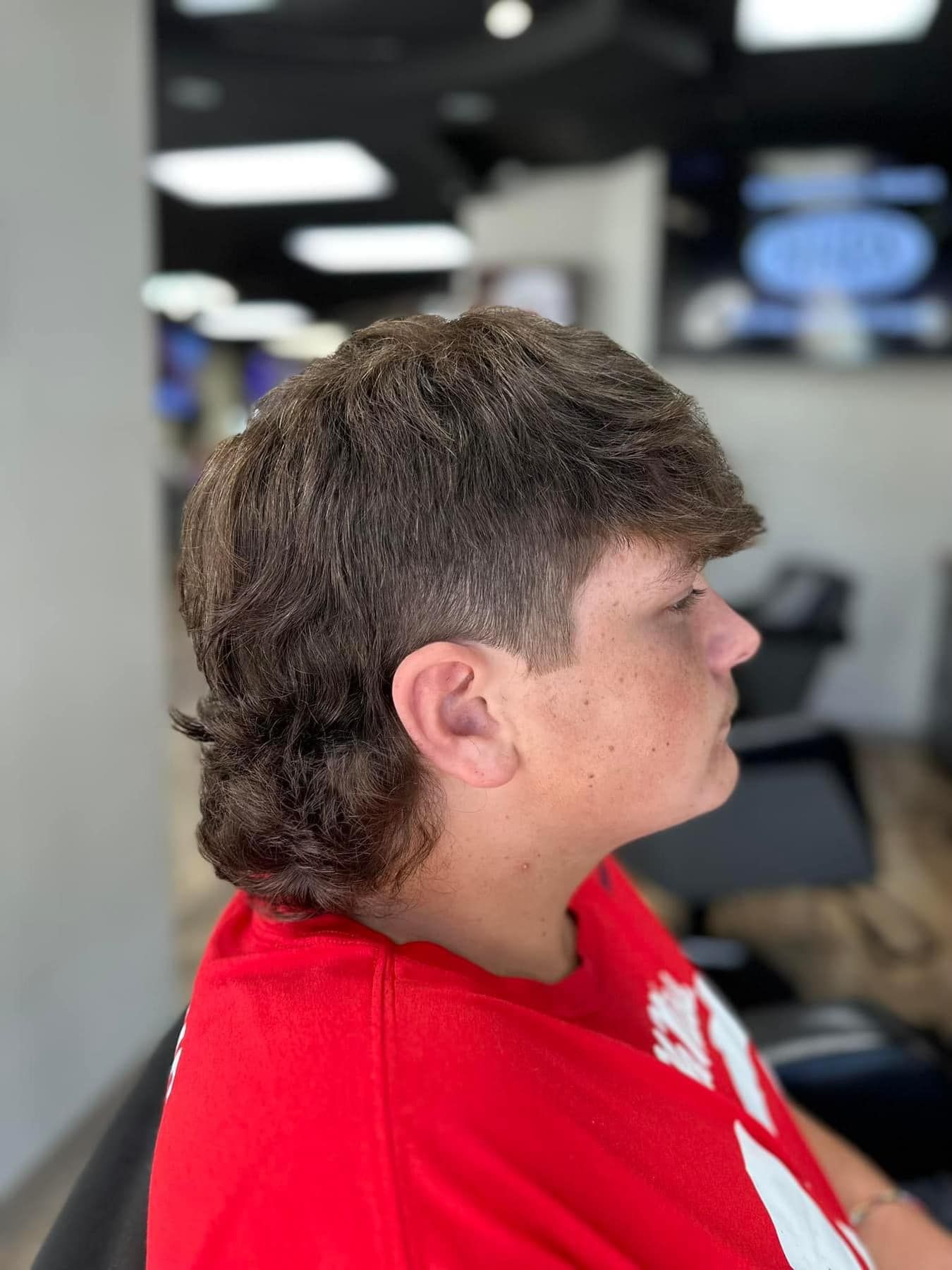 Side profile of a person with a mullet haircut wearing a red shirt in a blurred indoor setting