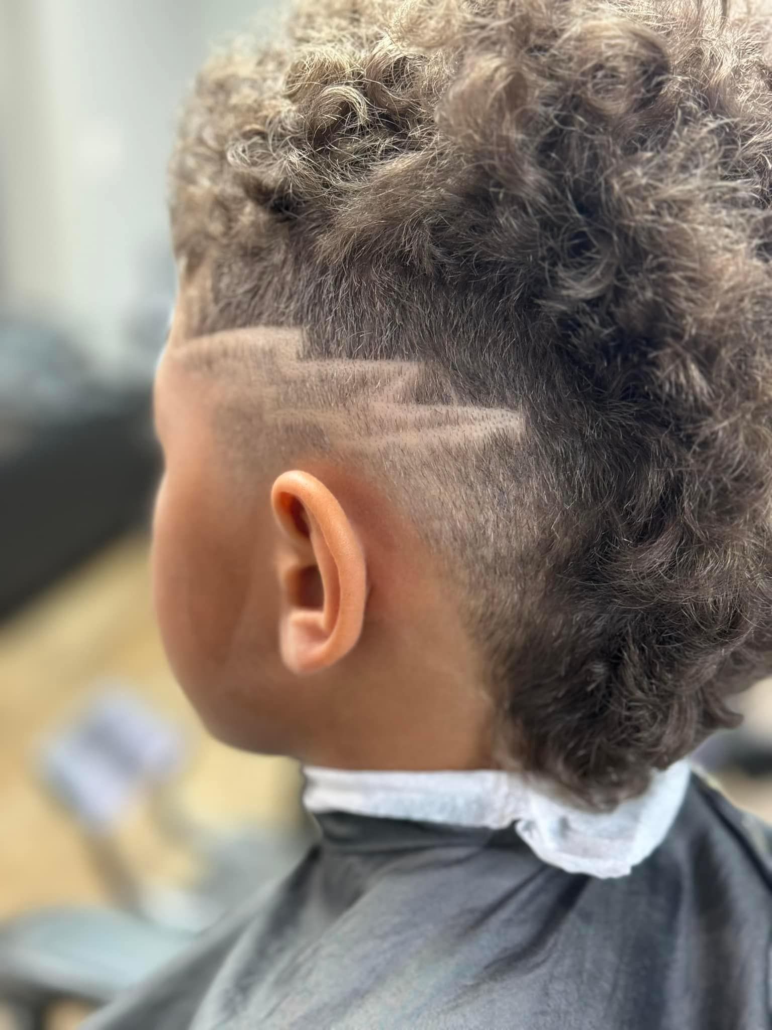 Child with curly hair and faded side cut, seated in a barber chair with a white neck strip.