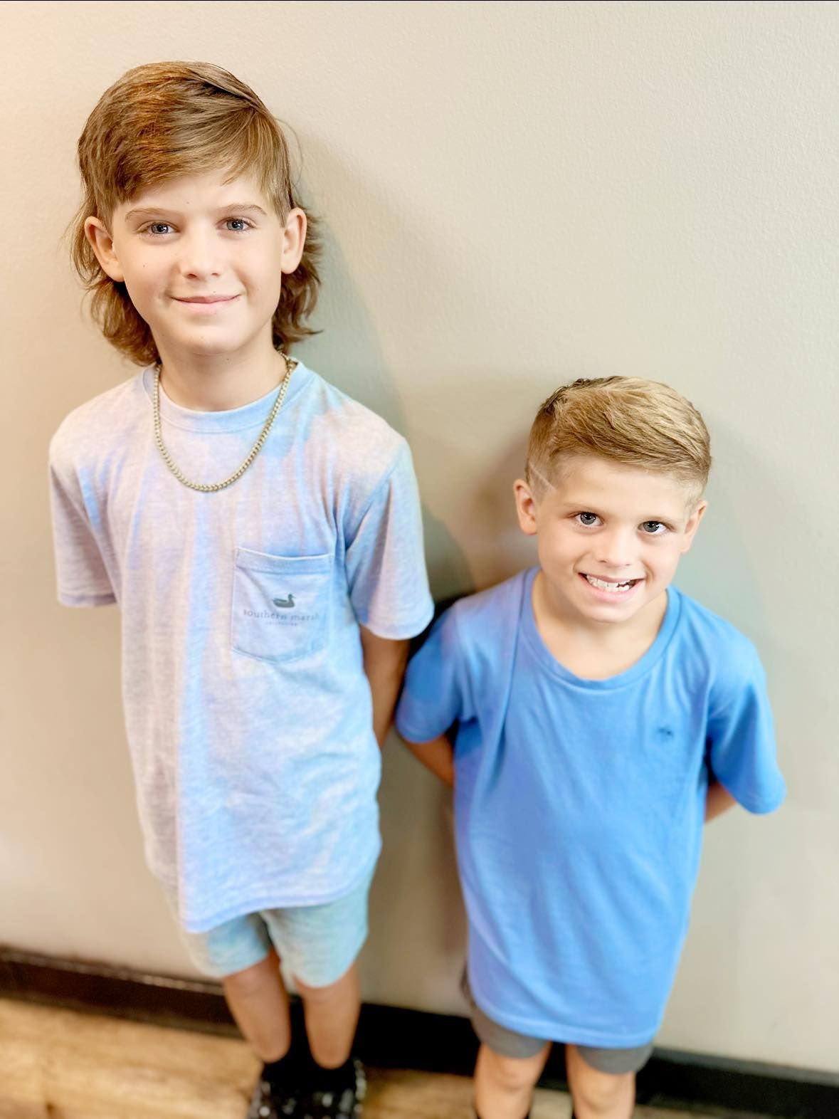 Two children standing against a wall, smiling, wearing light blue shirts and shorts.