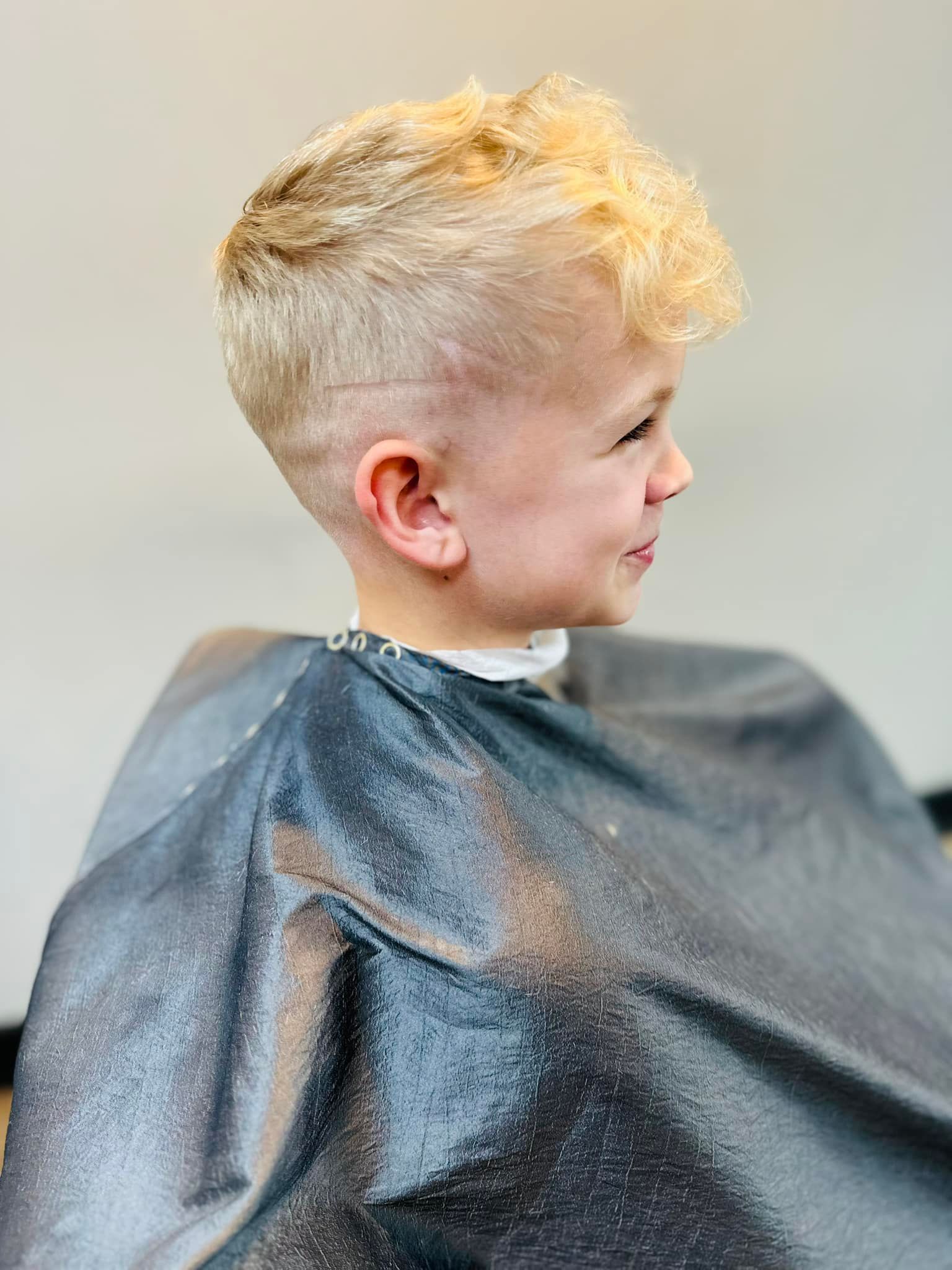 Child with short blond hair in a silver salon cape, shown in profile at a haircut.