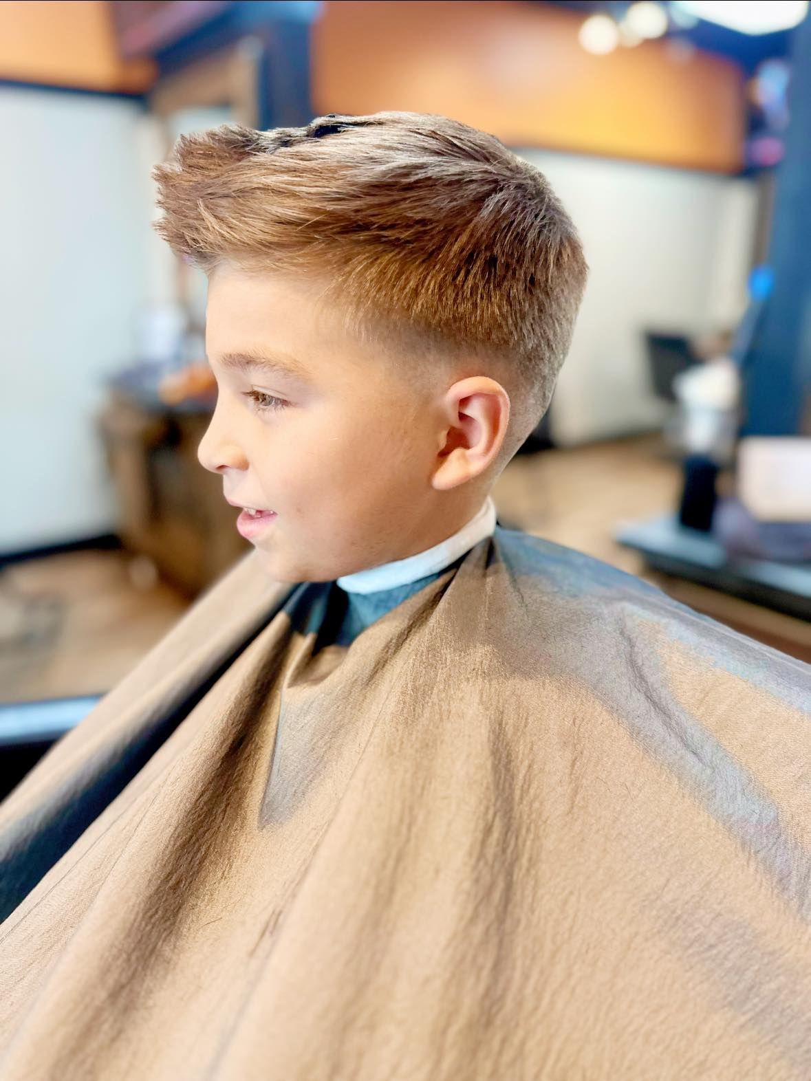 Child getting a haircut in a bright barbershop, wearing a beige cape and looking to the side.