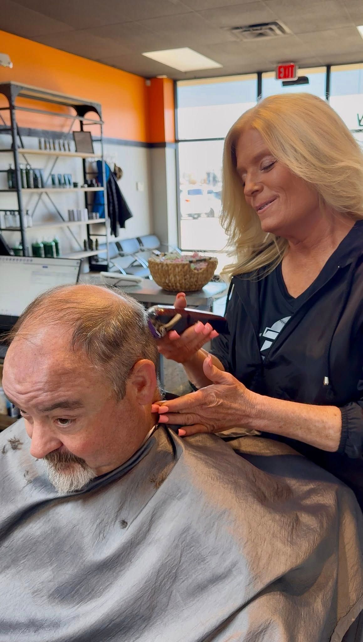 Hairdresser using clippers to trim a client's hair in a salon. Client is wearing a cape, and the hairdresser has blonde hair.