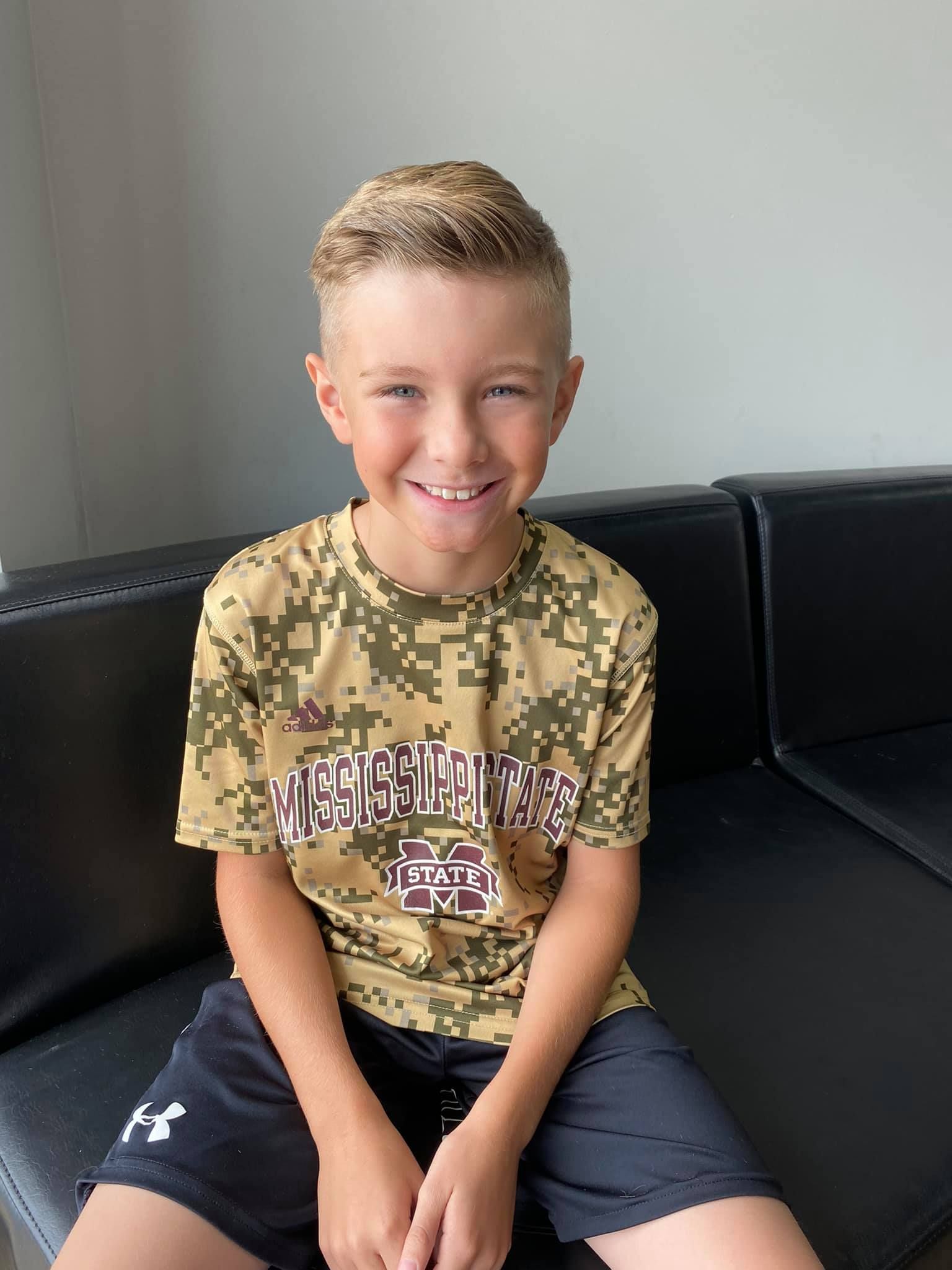 Smiling child sitting on a black couch, wearing a camouflage shirt and dark shorts.