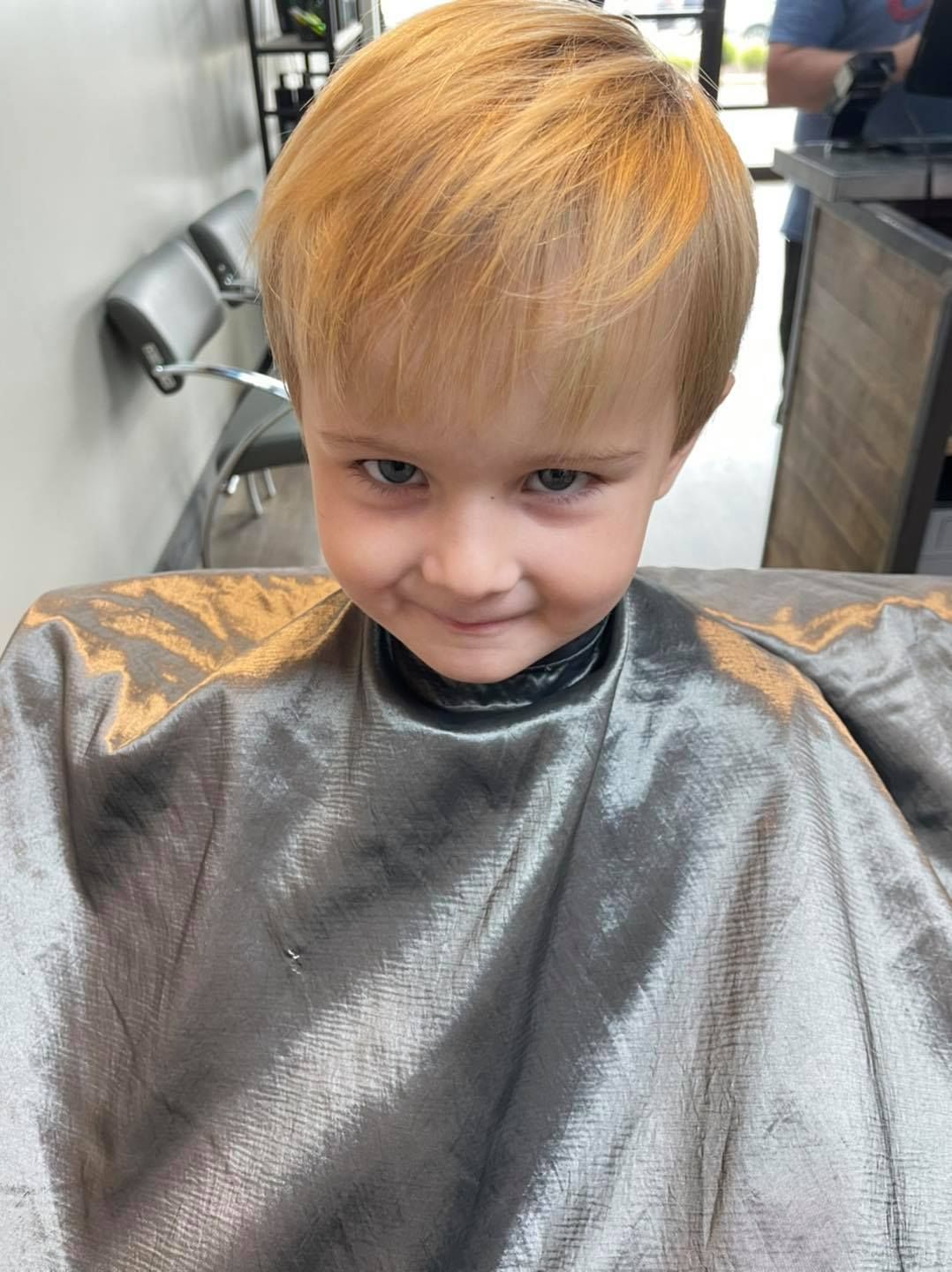 Child with short blond hair sitting in a salon cape, getting a haircut.