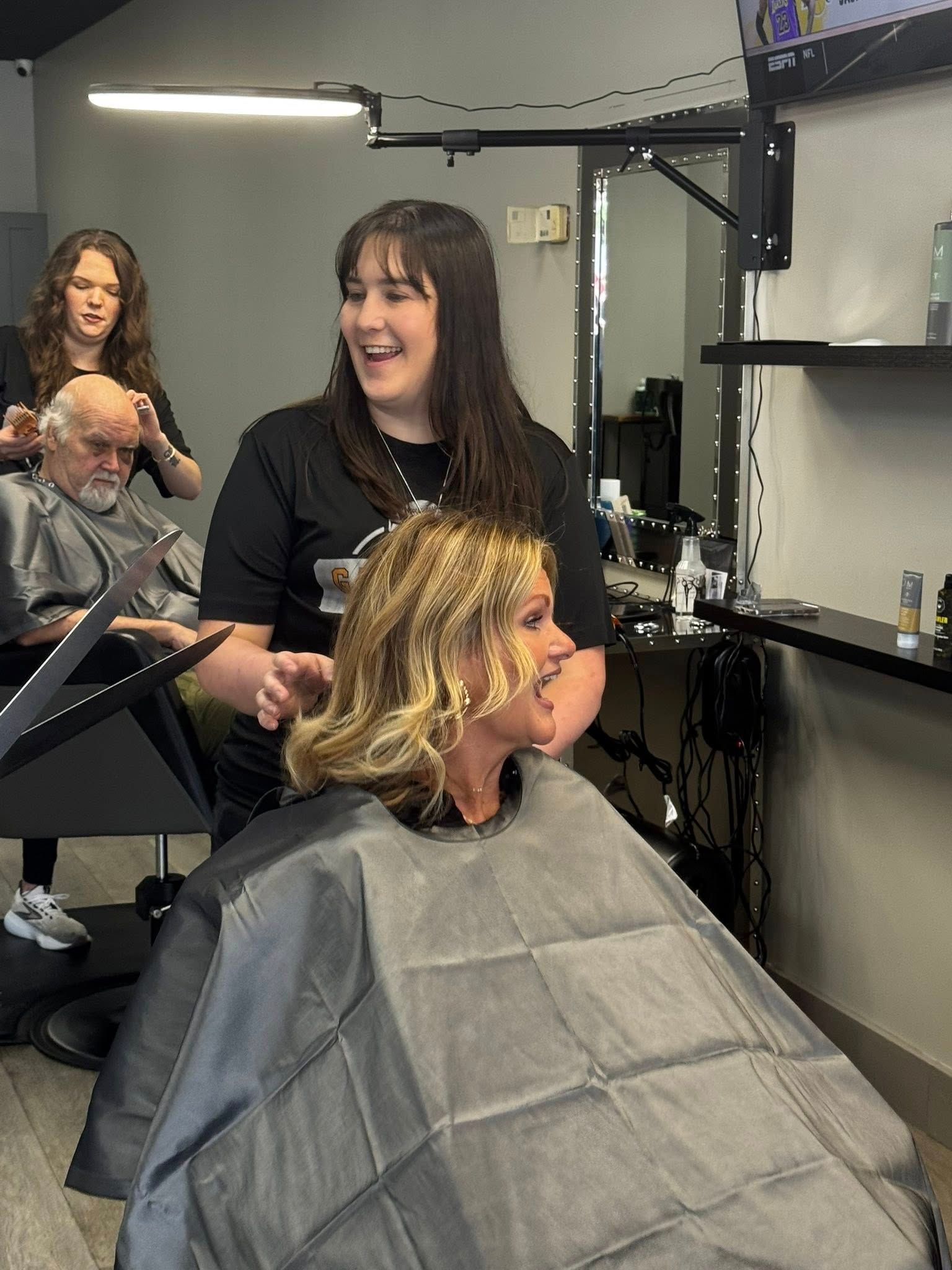 Salon scene: Two stylists cut clients' hair. One stylist smiles, blonde client in chair. Gray cape.