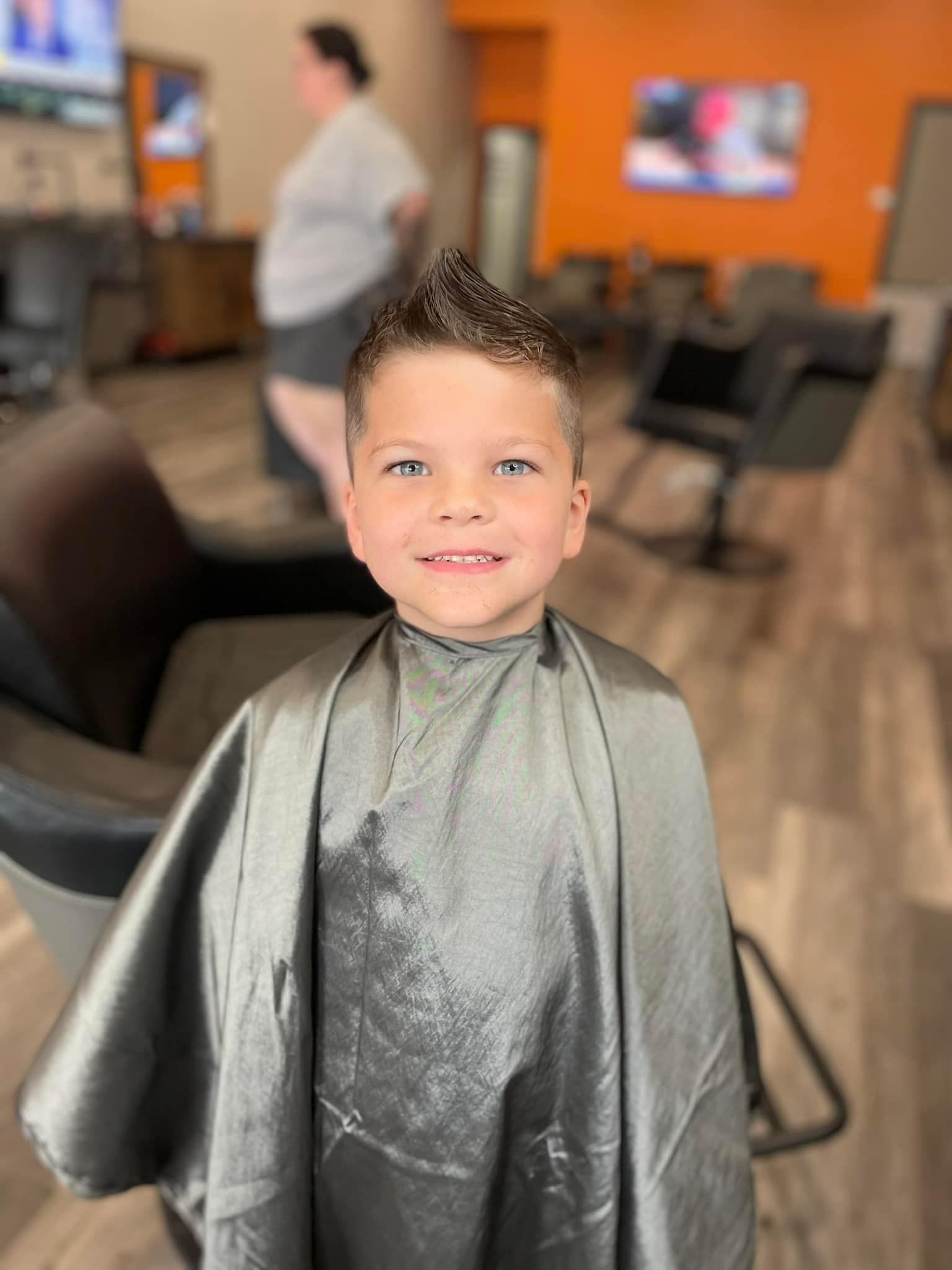 Child smiling in a barber chair with a silver haircut cape, inside a barbershop.