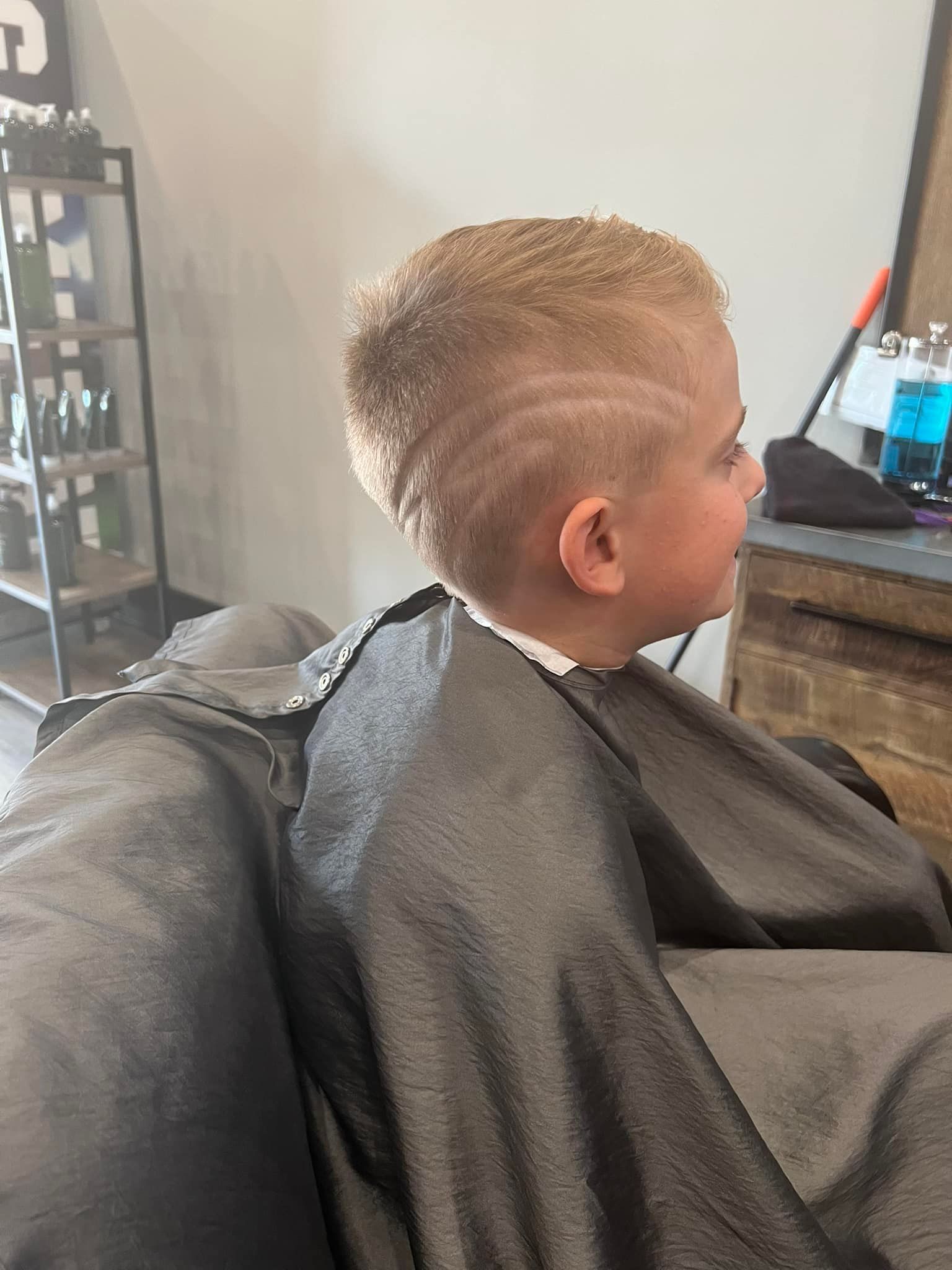 Child with short blond hair sitting in a salon chair under a black cape, facing a mirror.