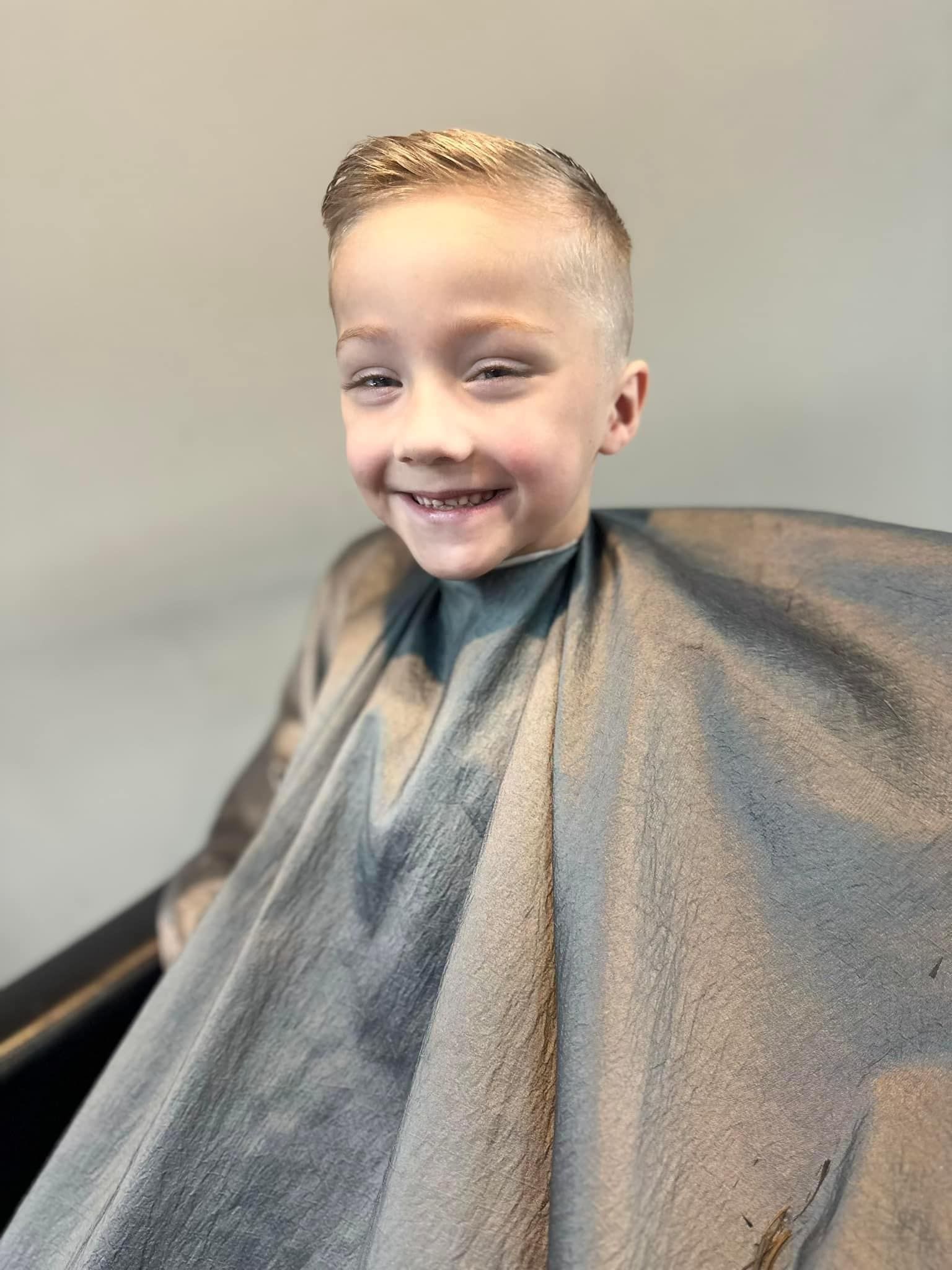 Smiling child seated in a salon chair, draped in a gray haircut cape.