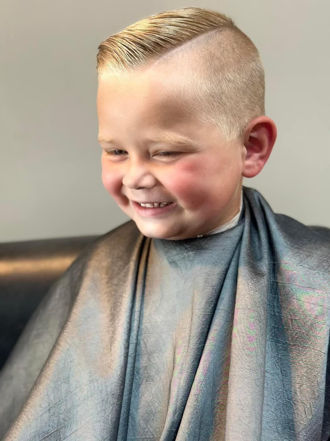 Smiling child with a fresh short haircut, wearing a gray barber cape in a salon chair