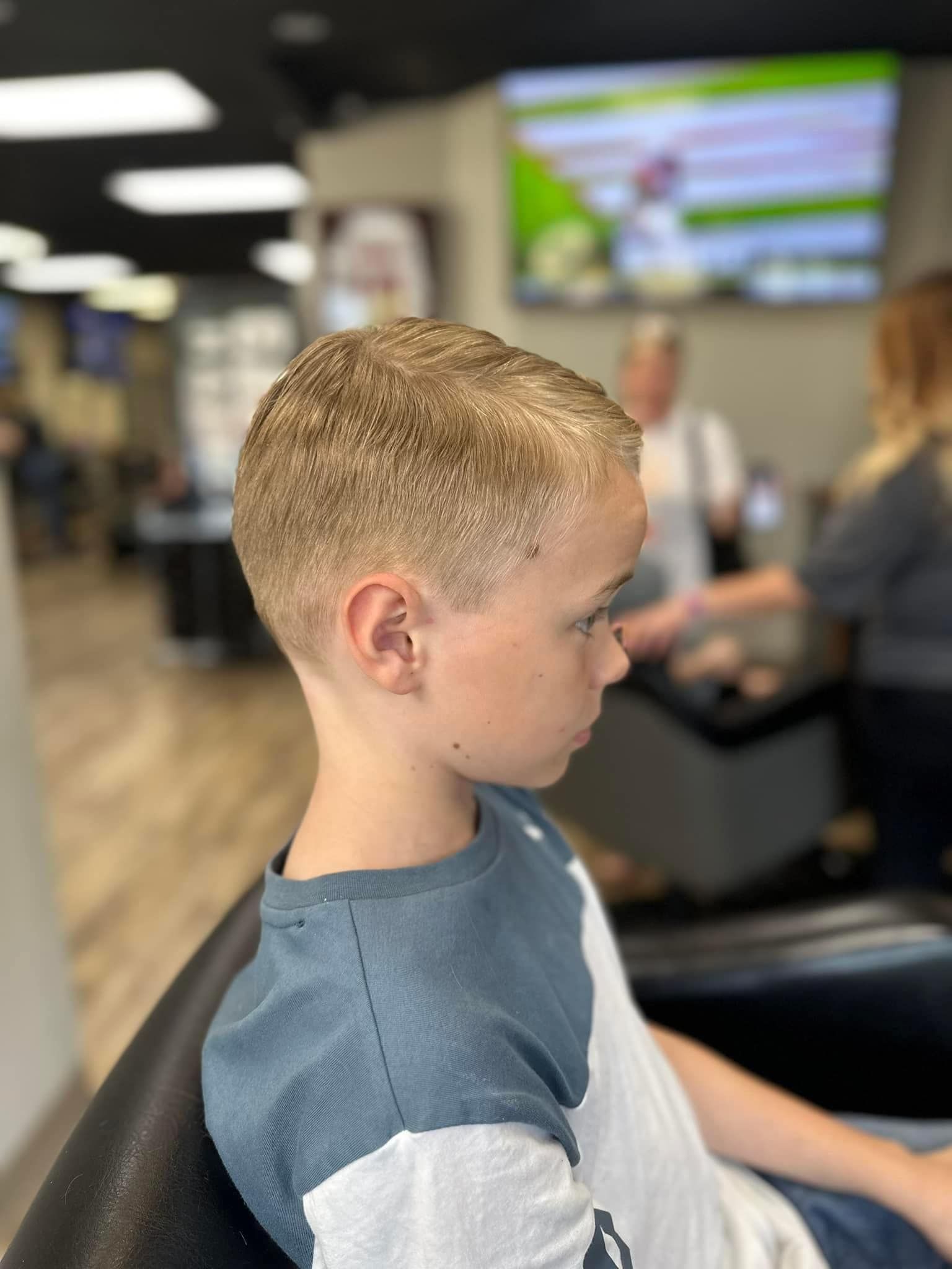 Boy with short blond hair sitting indoors in a waiting area, facing right, with a blurred TV in the background