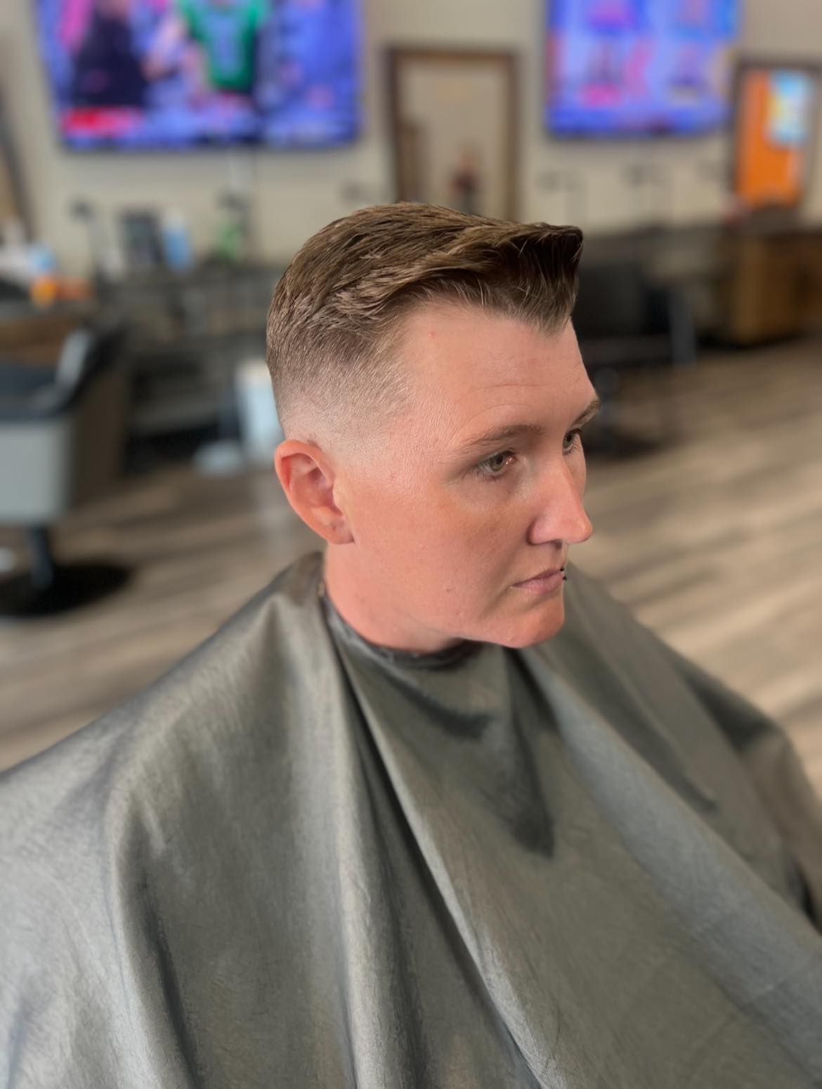 Person with a short fade haircut in a barber cape, seated in a modern barbershop interior.
