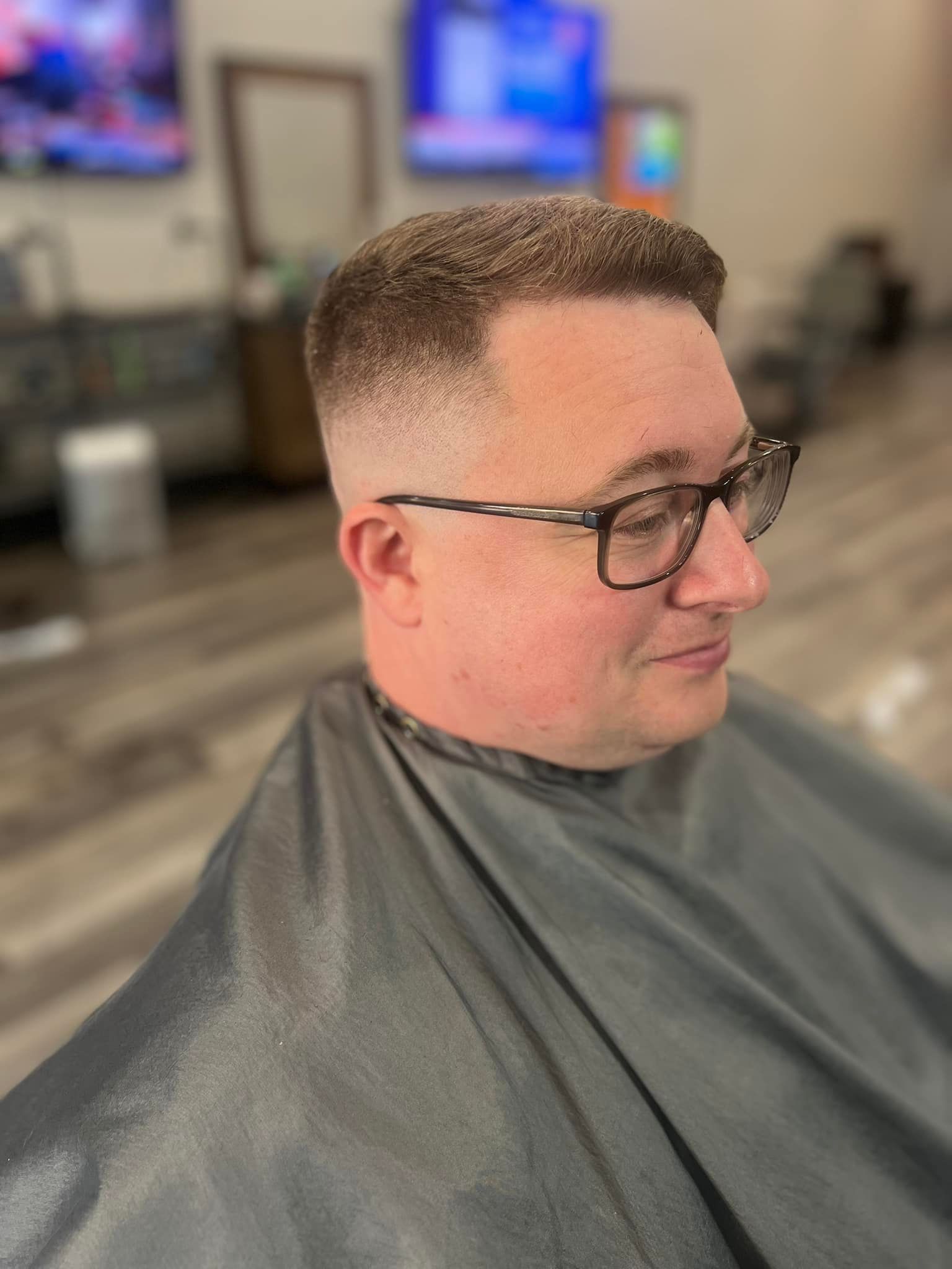 Man with glasses and a fresh fade haircut sitting in a barbershop cape, looking right.