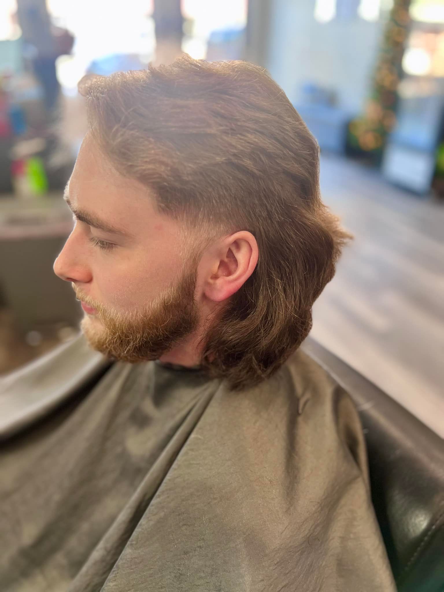Profile view of a man with a mullet and beard getting a haircut, wearing a salon cape