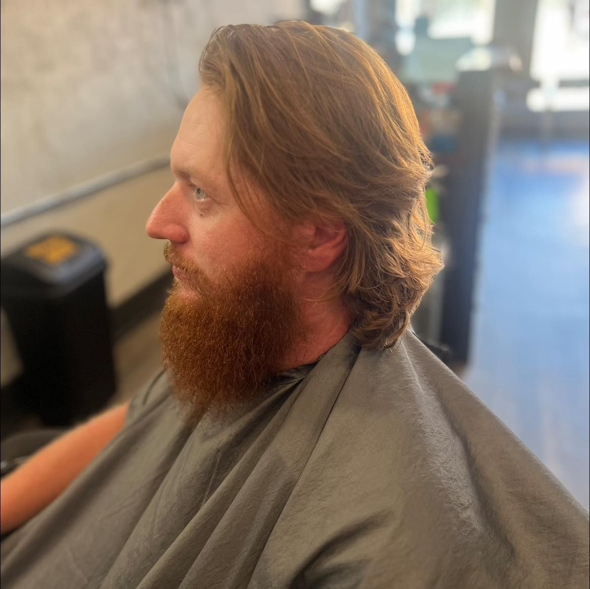 Man with a red beard seated in a salon chair, wearing a gray cape, shown in profile.