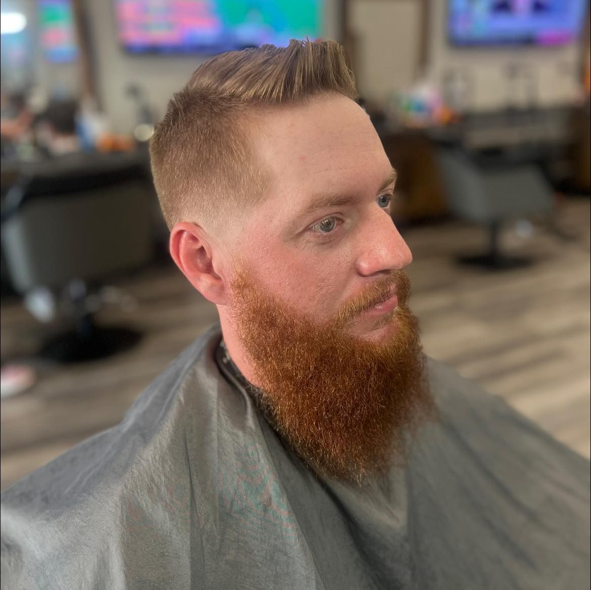 Side profile of a man with a red beard in a barbershop chair, wearing a gray cape