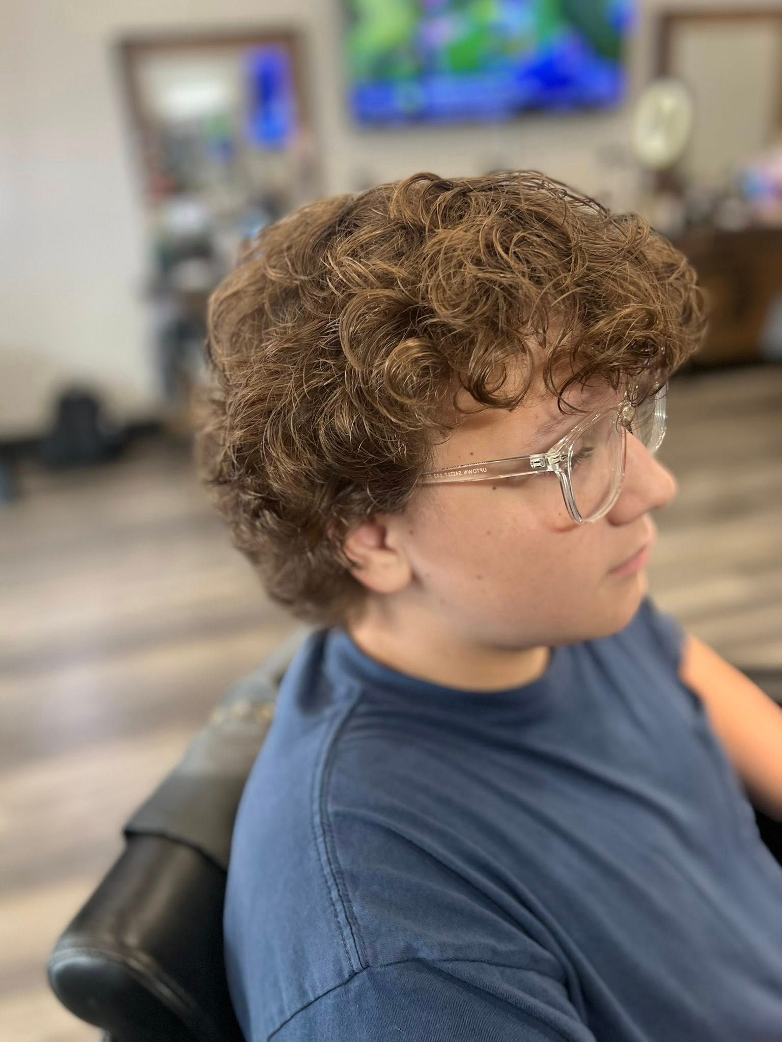 Person with curly hair and glasses sitting in a chair in a blurred indoor setting.