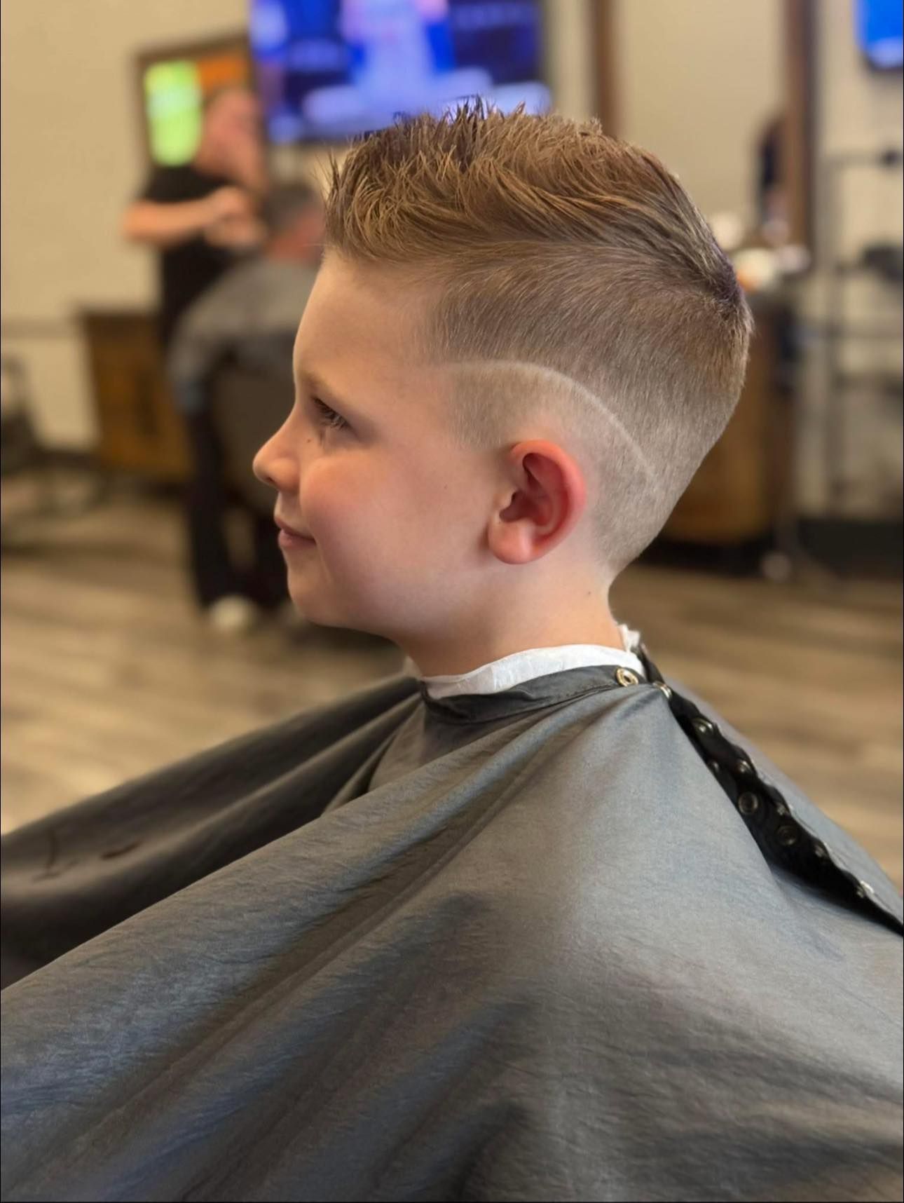 Child with a short fade haircut sitting in a barber chair, wearing a black cape in a barbershop.