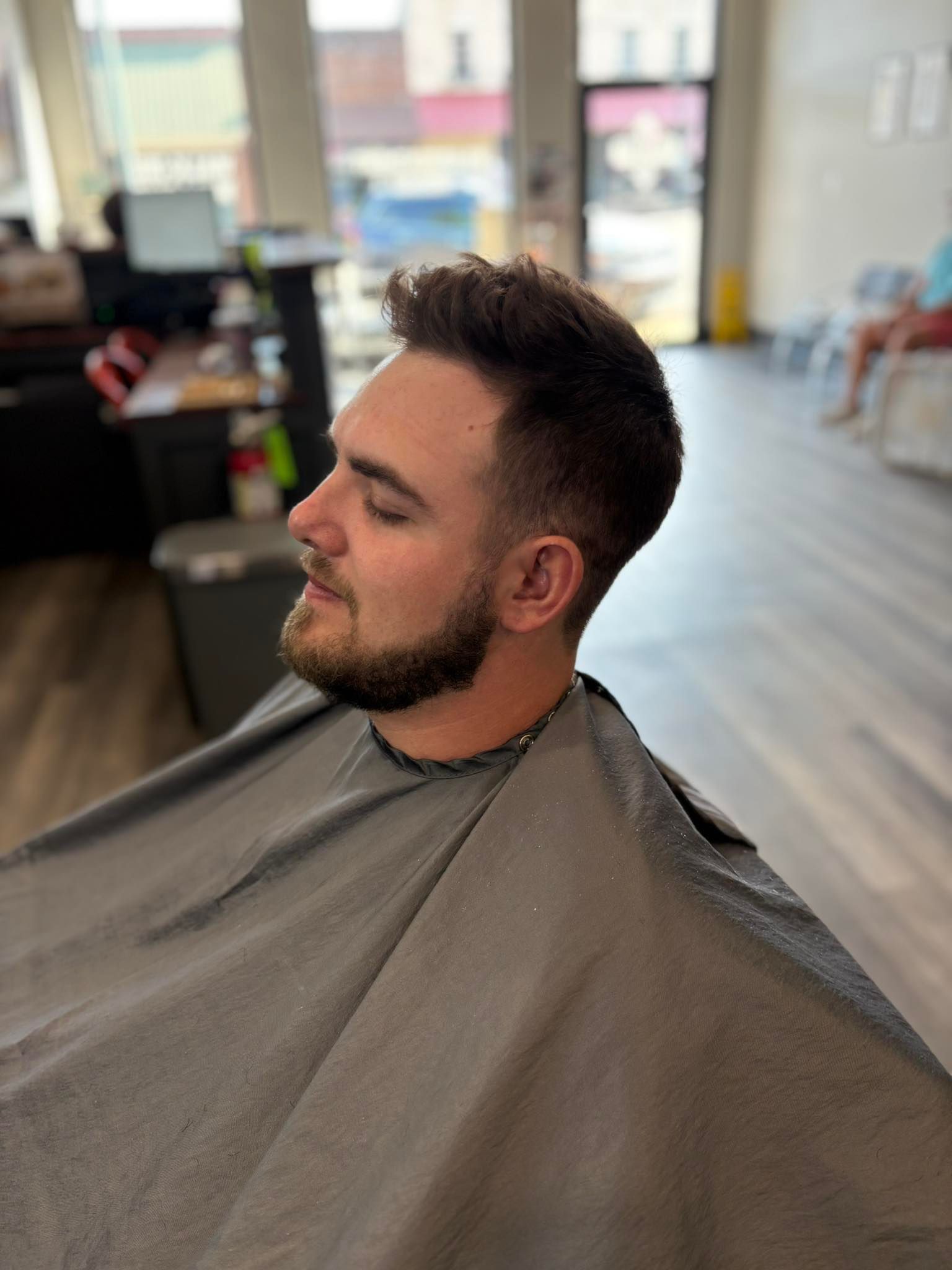Man with short dark hair and beard sitting in a salon cape during a haircut in a bright barbershop