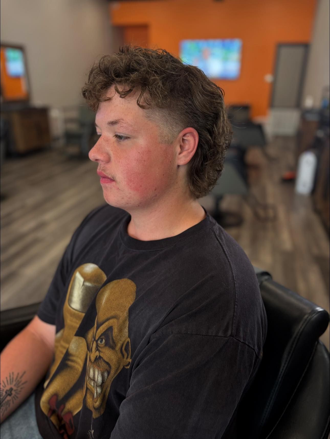 Person with curly hair in a black graphic T-shirt seated indoors, shown in side profile with a blurred background