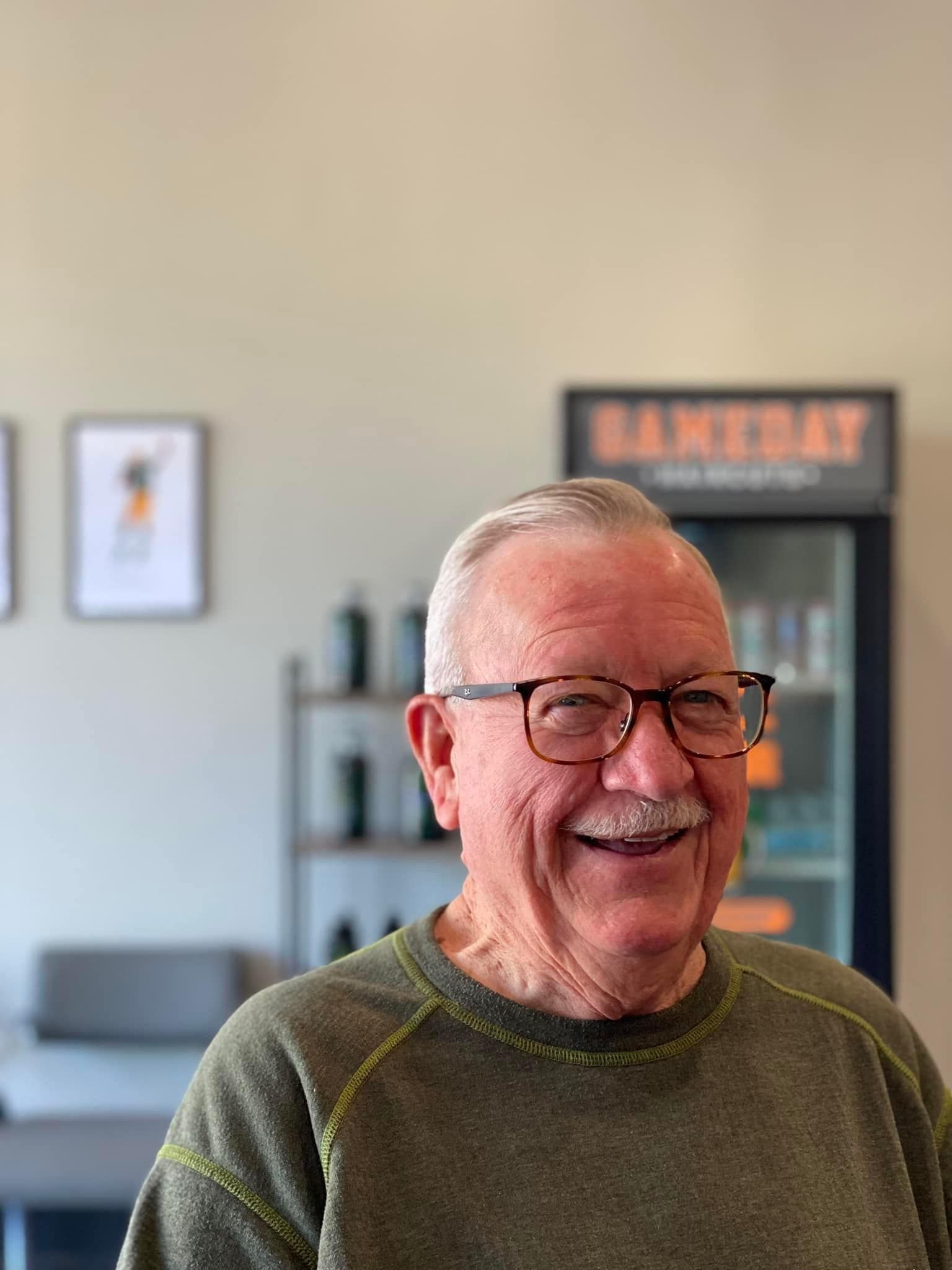 Smiling man in glasses and an olive sweater standing indoors, with a blurred shelf and sign behind him.