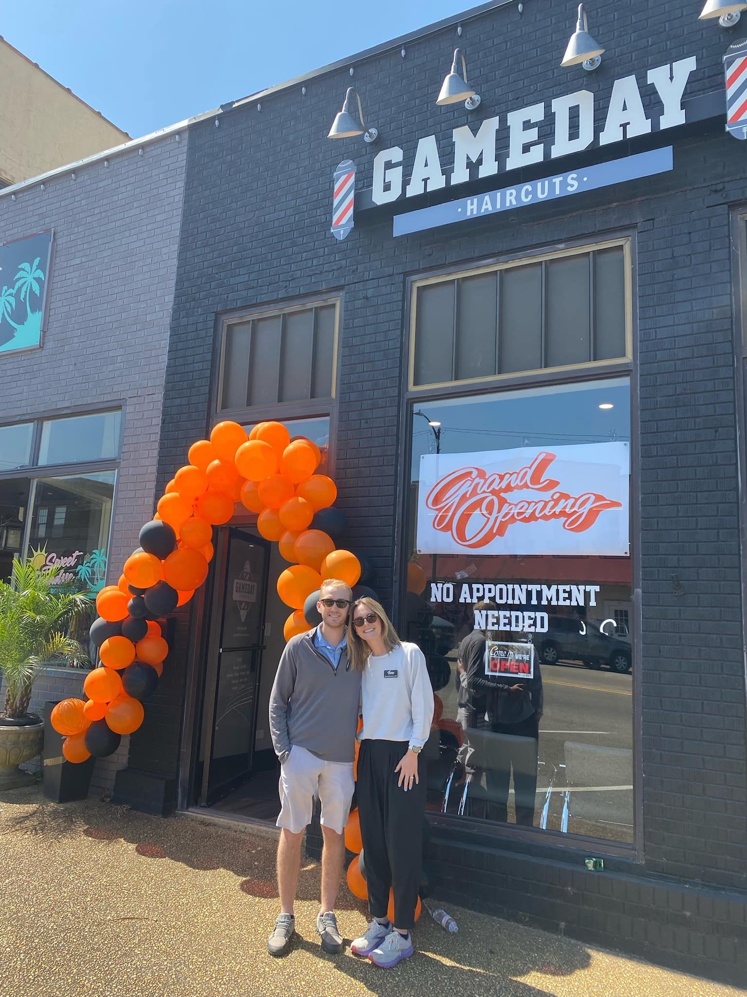 A couple stands in front of "Game Day Haircuts" shop, decorated with orange and black balloons.