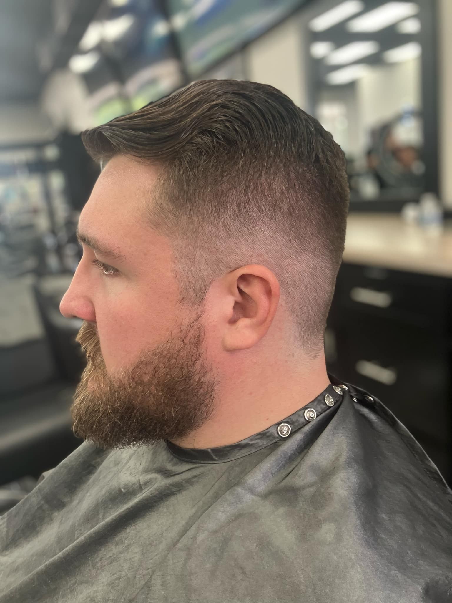 Side profile of a bearded person with a short fade haircut, wearing a barber cape in a salon chair.