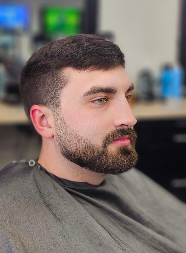 Profile view of a bearded man with short dark hair in a barber chair, wearing a gray cape
