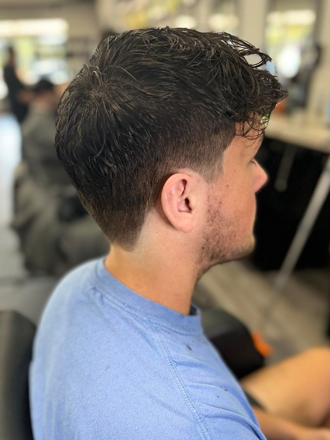 Side view of a man with dark curly hair and faded haircut, wearing a light blue shirt in a barbershop.