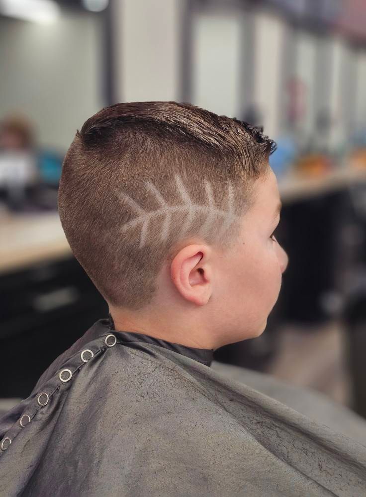 Child with a short haircut and shaved zigzag design, seated in a barber chair with a gray cape.