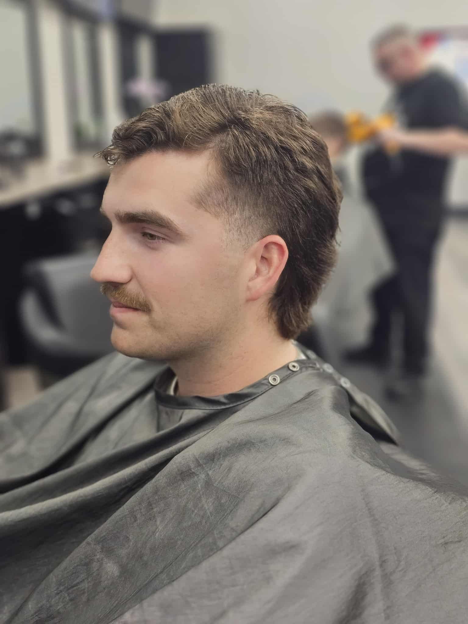 Person with short curly hair seated in a barber chair wearing a dark cape, in a salon.