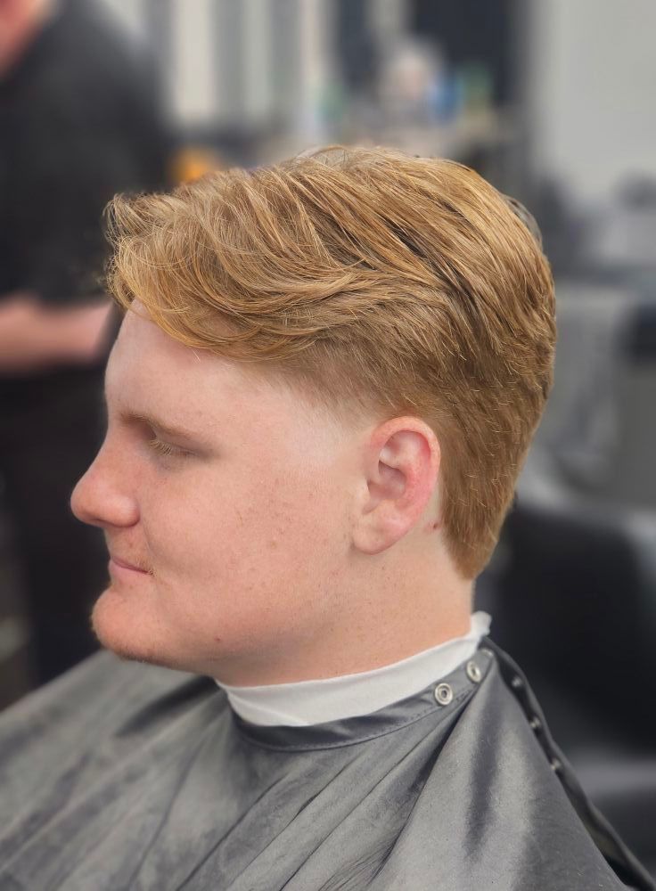 Side profile of a person with short blond hair getting a haircut, wearing a gray barber cape.