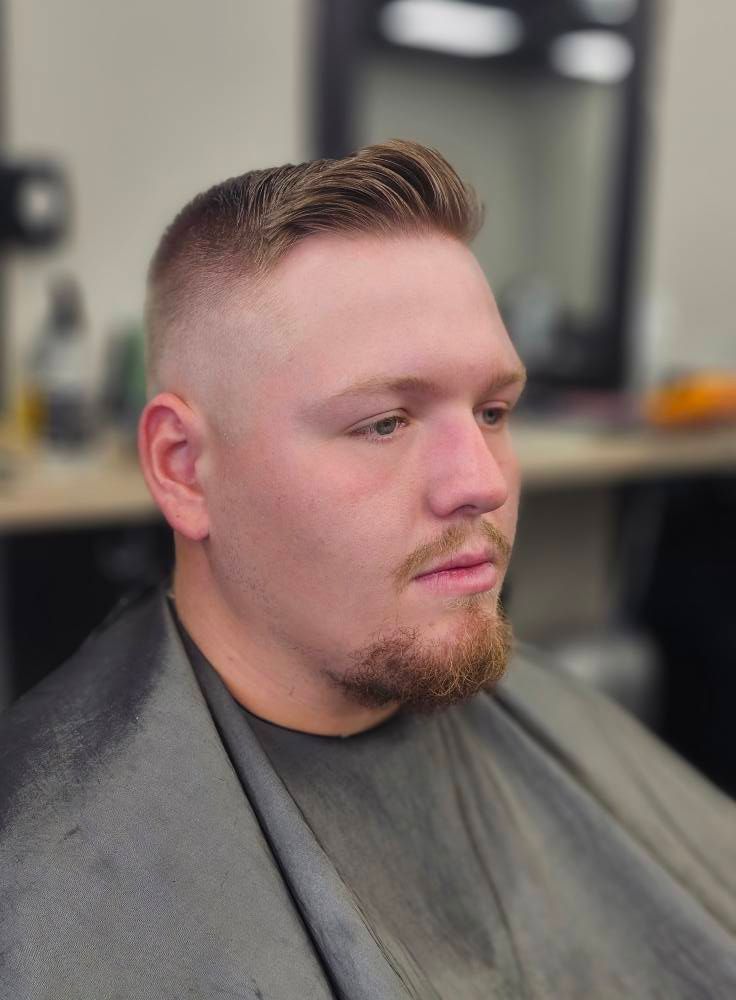 Man with short faded haircut and trimmed beard in a salon chair, wearing a gray cape, looking to the side