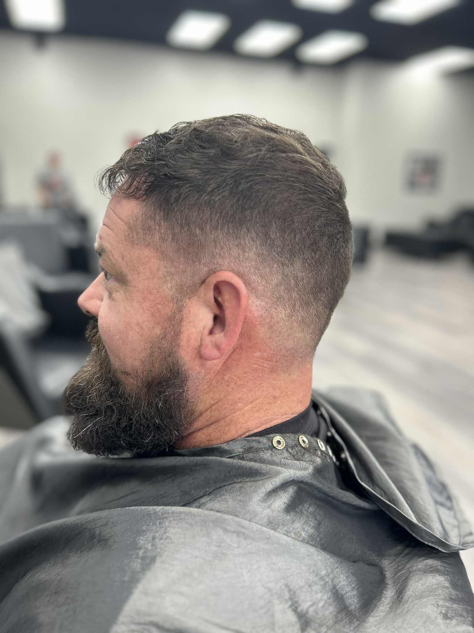 Close-up of a man with a short fade haircut and full beard, seated in a barber chair in a salon.