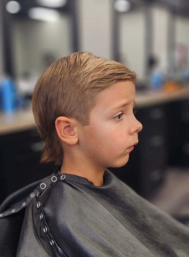 Child in a black salon cape, side profile, getting a haircut in a blurred barbershop.