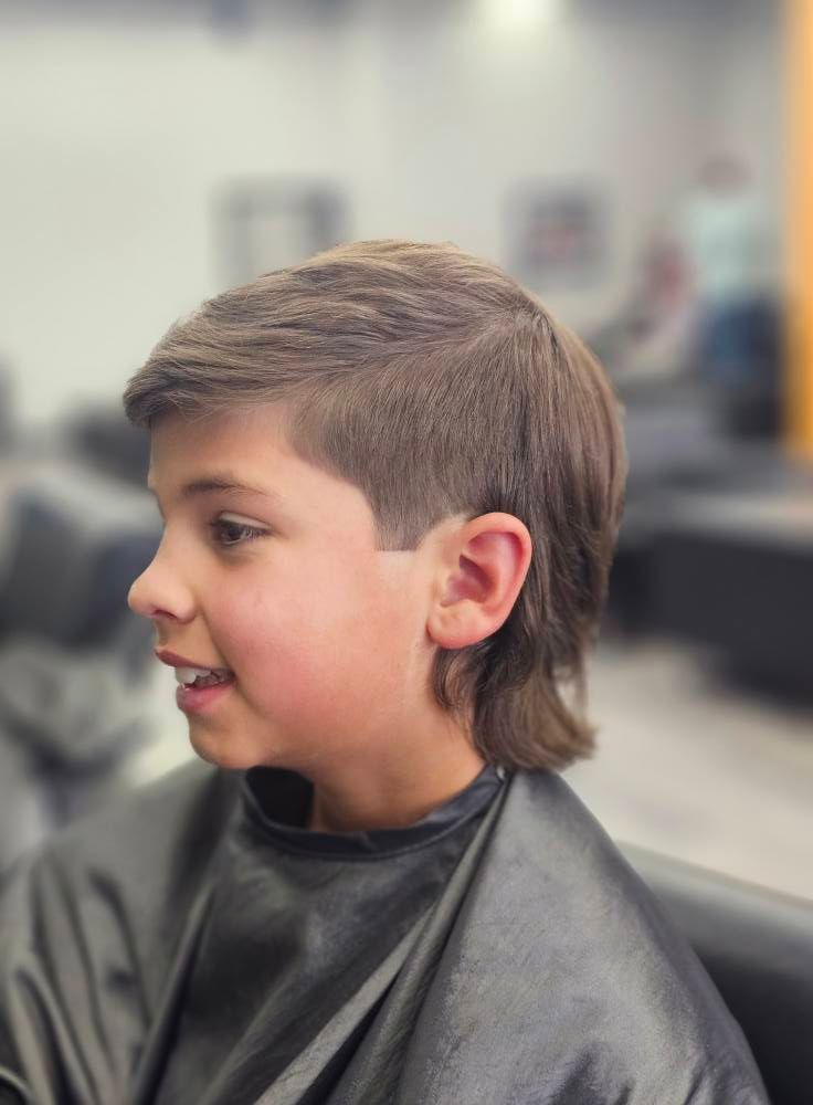 Profile view of a child with a mullet haircut in a salon chair, wearing a black cape