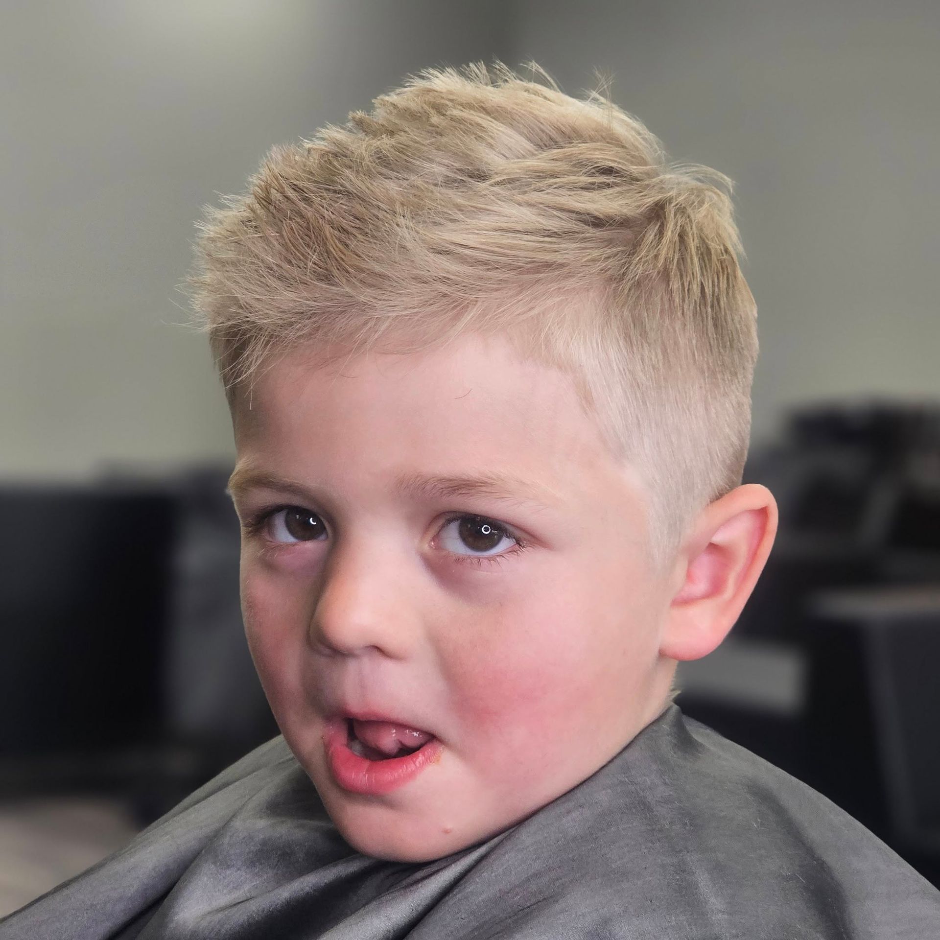 Close-up of a blond child with a tongue out, wearing a gray cape, against a blurred indoor background