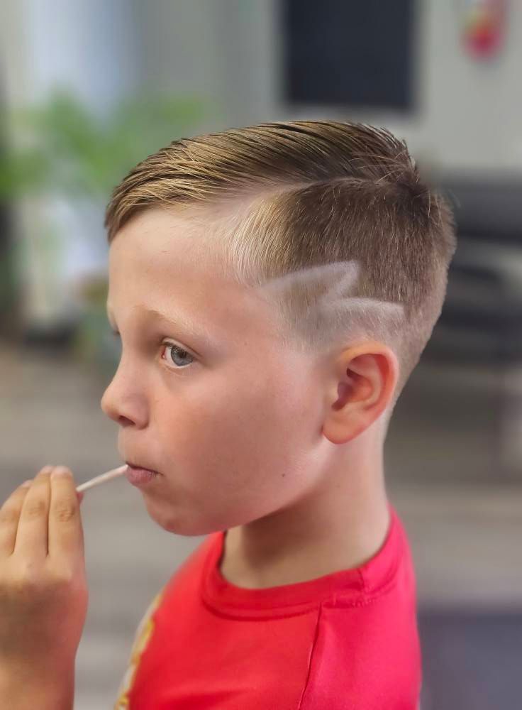 Child with a side haircut blowing bubbles outdoors, wearing a red shirt.
