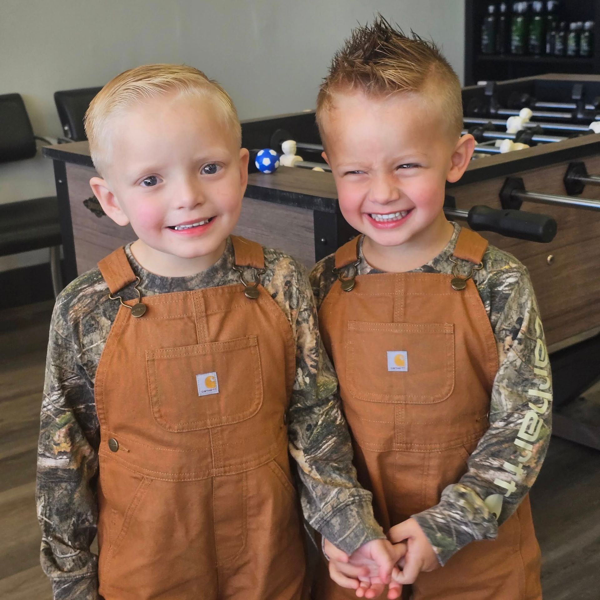 Two smiling children in brown overalls and camouflage shirts standing indoors with hands clasped.