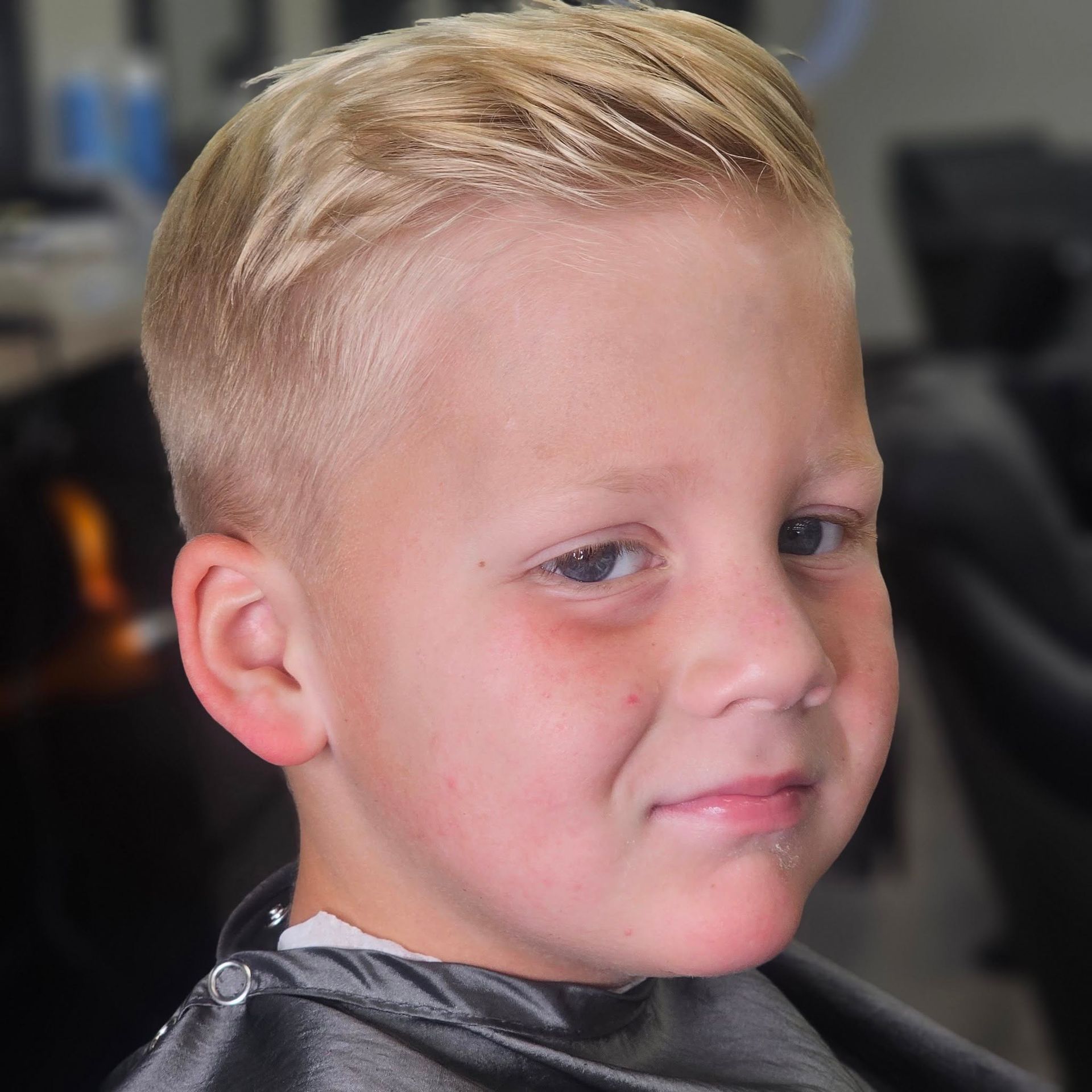Close-up portrait of a young child with short blond hair, wearing a dark cape, against a blurred indoor background