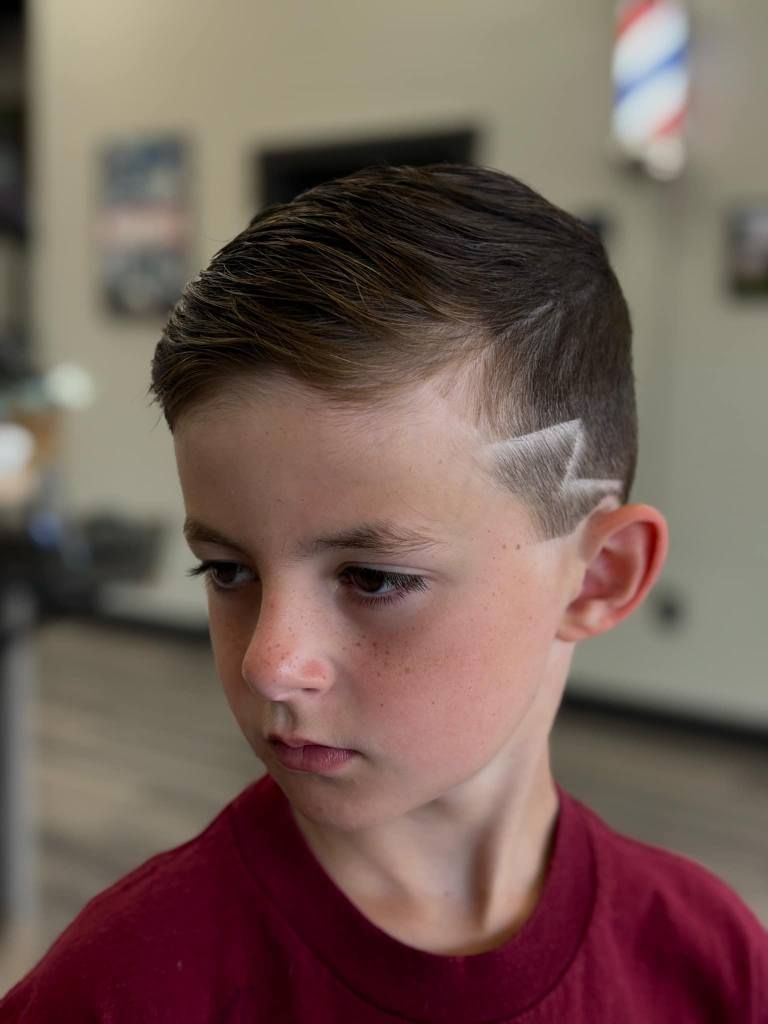 Boy with a side-fade haircut and shaved lightning design, wearing a maroon shirt, looking left indoors