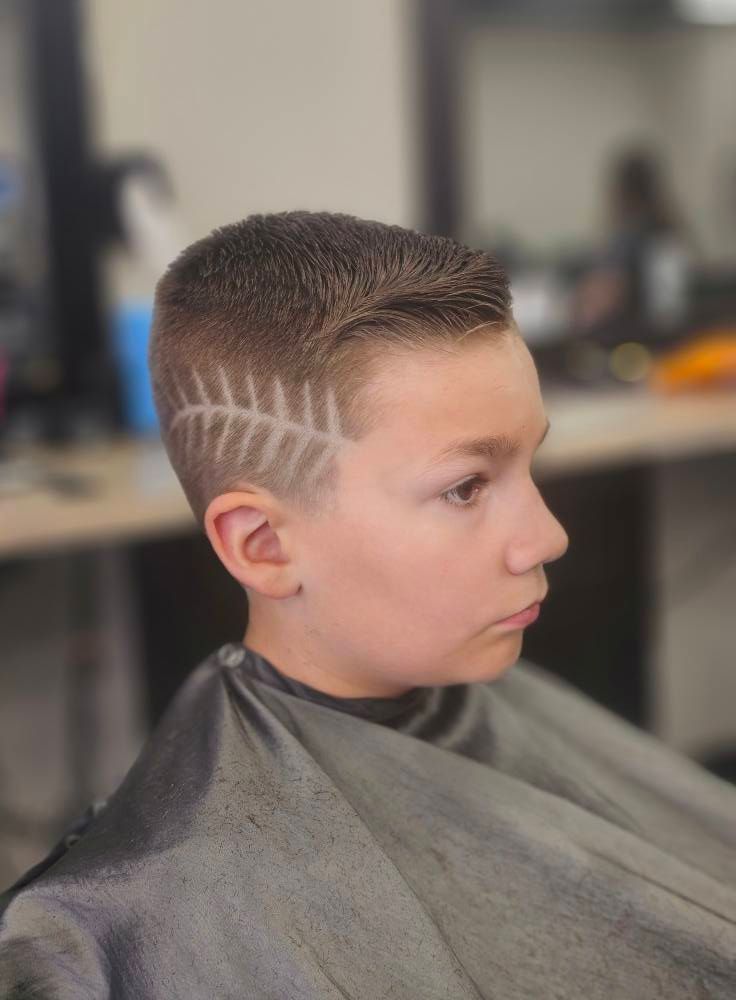 Boy in a barbershop with a fresh short haircut and shaved design on the side, wearing a gray cape.