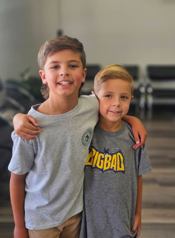 Two boys standing together, smiling, with one arm around the other in a casual indoor setting.