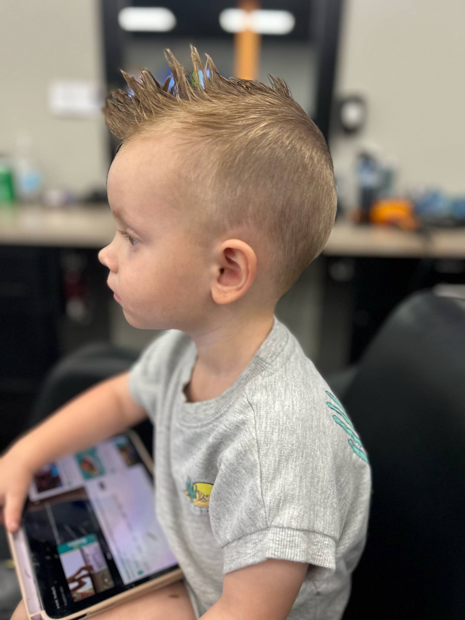 Profile of a child with a spiky blond mohawk, holding a tablet in a salon chair