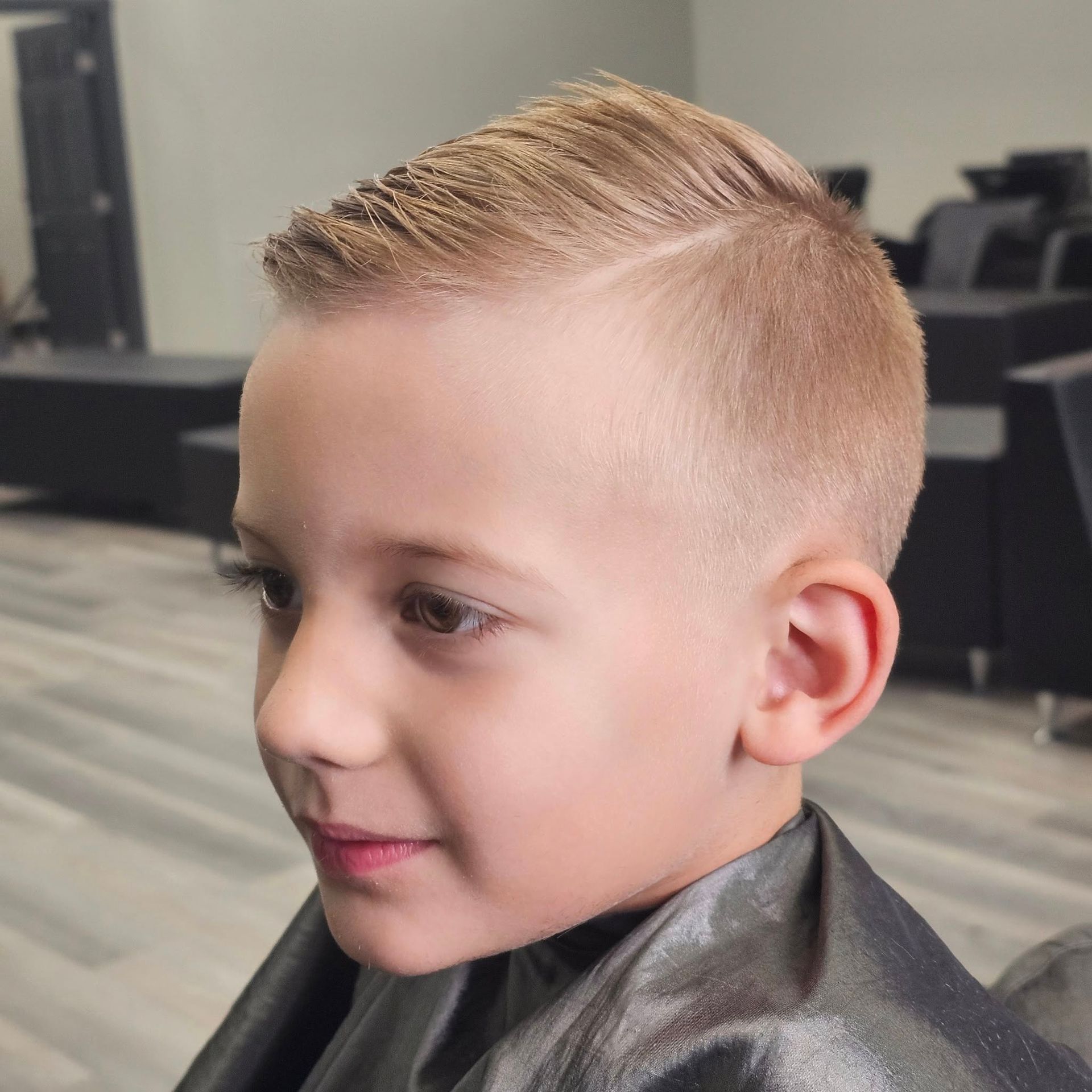 Young child with a neat blond haircut, seated in a salon chair, looking to the side.