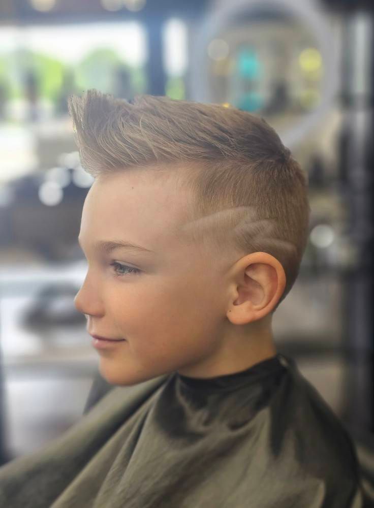 Child with a spiked haircut in a barber chair, side profile, in a salon with blurred background.