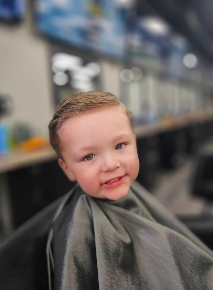 Child with a fresh haircut, smiling in a barber chair with a dark cape draped around them.